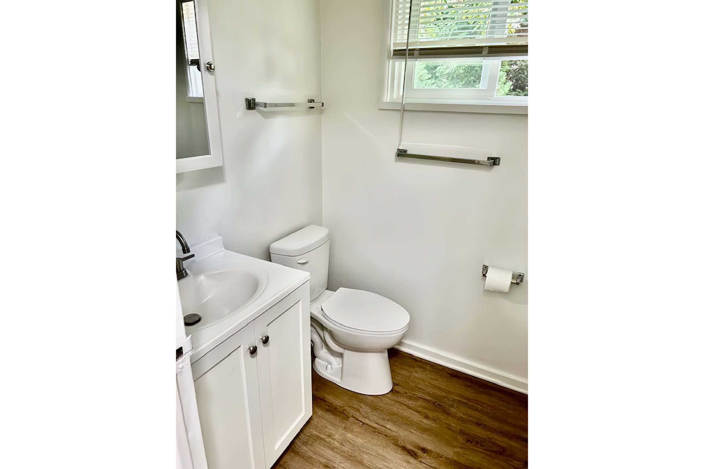 A bright, modern bathroom featuring a white sink with a cabinet underneath, a white toilet, and a roll of toilet paper on a holder. Light-colored walls and a window allow natural light to brighten the space, complemented by wooden flooring and polished metal towel racks.