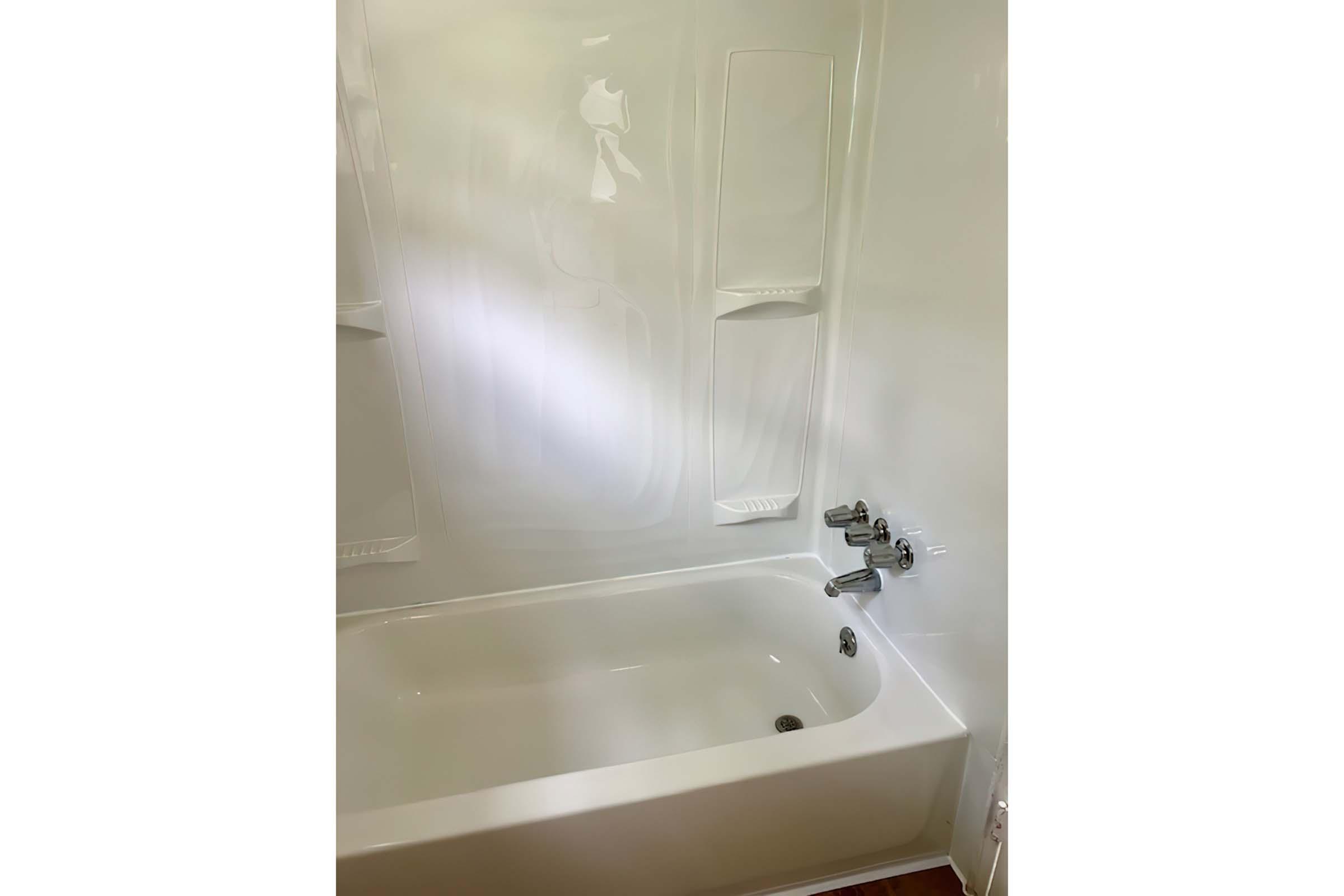 A clean, white bathtub with a simple design. The tub features a smooth, glossy finish and is surrounded by plain walls. There are three faucet handles visible on the side wall, and two built-in shelves are located directly behind the tub for storage. The overall atmosphere is fresh and uncluttered.