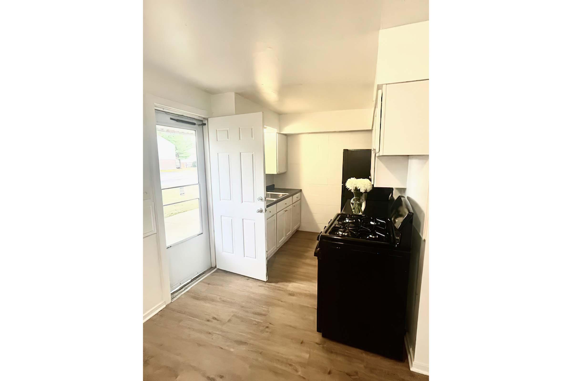 A modern kitchen featuring white cabinets, a black stove, and an open layout. There is a large window with a door leading outside, providing natural light. The floor is covered with light-colored wood, creating a clean and spacious atmosphere. A vase with flowers adds a decorative touch to the space.