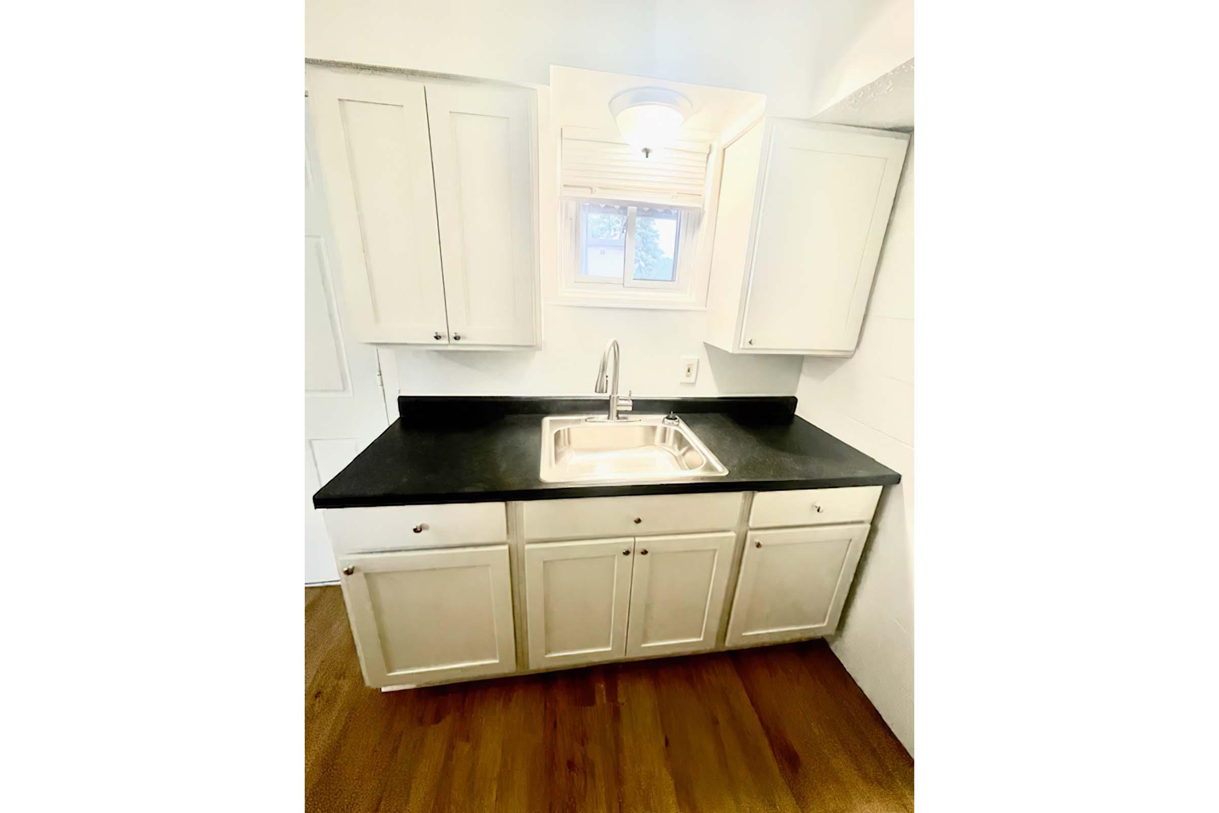 A small kitchen area featuring white cabinetry, a black countertop, and a stainless steel sink. Above the sink is a window allowing natural light to enter. The flooring is made of wood. The overall design is simple and functional.