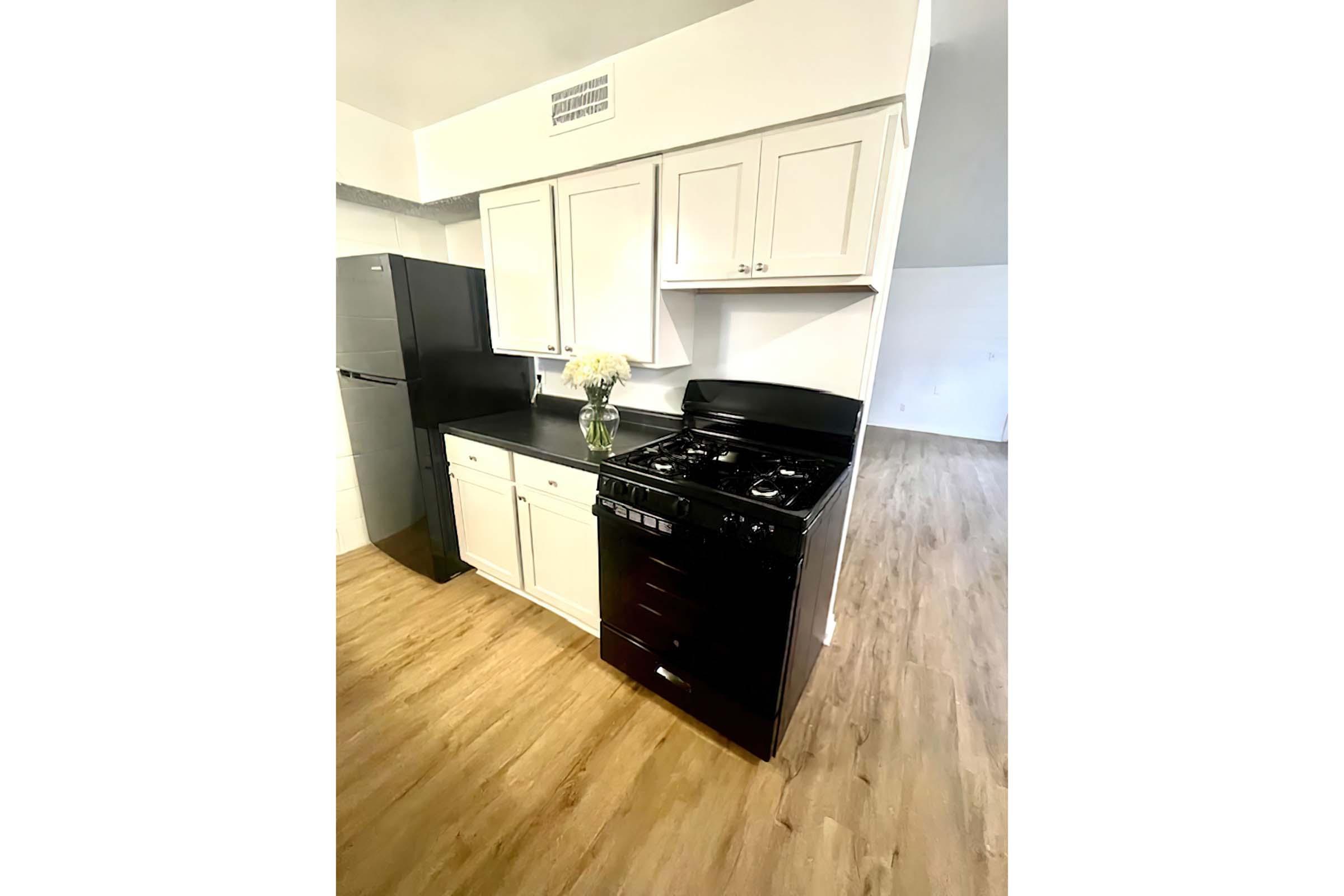 A modern kitchen featuring white cabinetry, a black stove, and a black refrigerator. The countertop is dark, and a vase of flowers adds a decorative touch. The flooring is light-colored wood, creating a bright and spacious atmosphere.