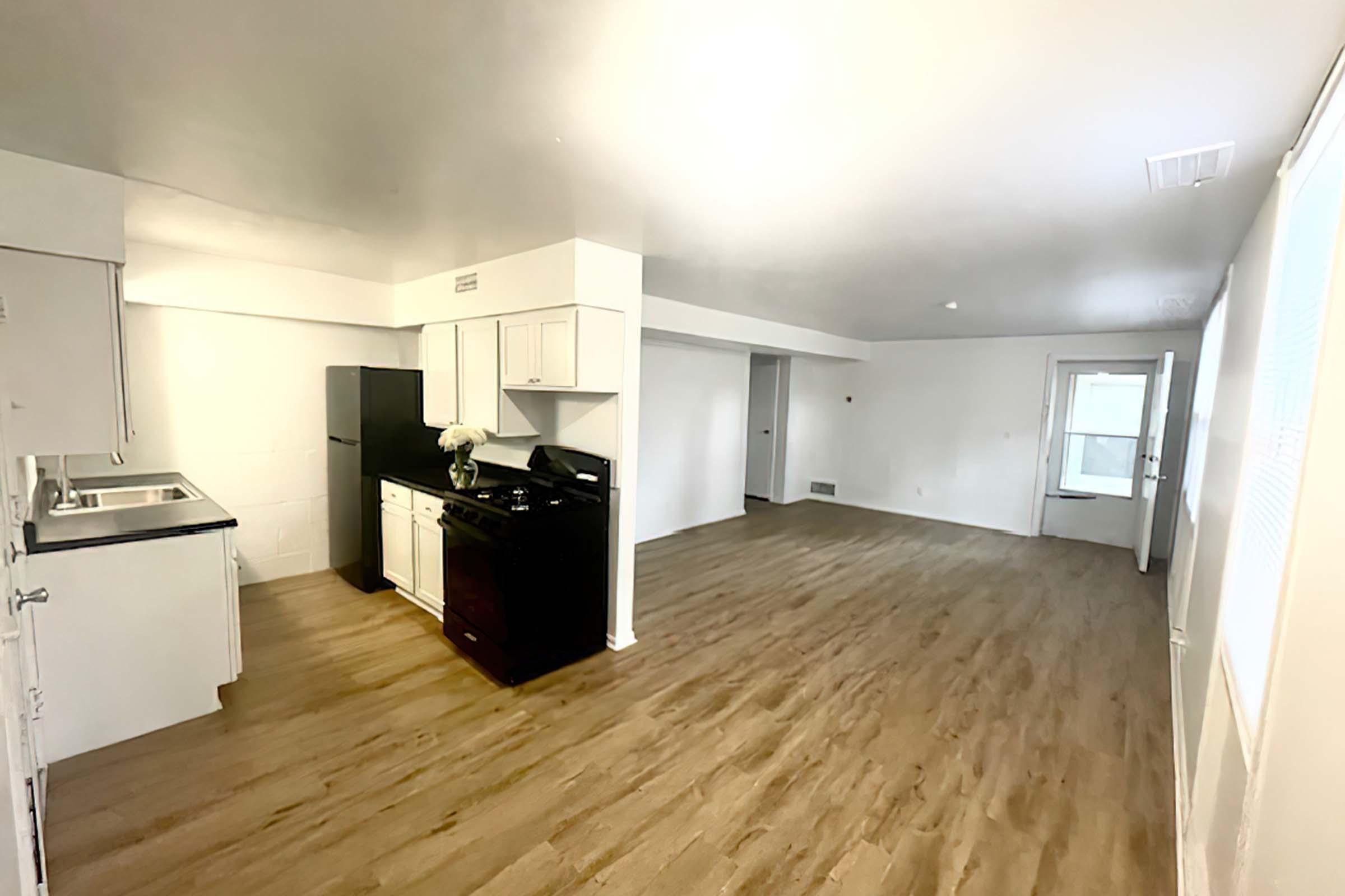 A spacious, bright interior of a modern kitchen and living area. The kitchen features white cabinets, a black refrigerator, and a black stove. Light wood flooring runs throughout the room, which has large windows allowing natural light and a door leading outside. The walls are painted a soft white, creating an open atmosphere.