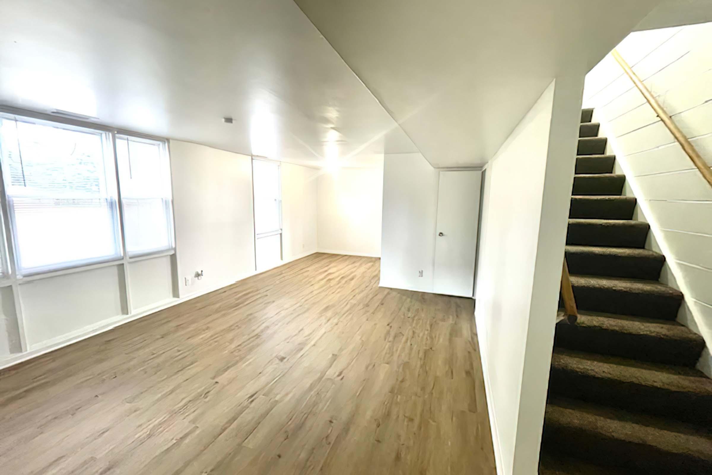 Spacious, empty room with light-colored walls and laminate flooring, featuring large windows that allow natural light. A staircase with wooden railing leads to an upper level. The room has a clean and minimalist aesthetic, suitable for various uses.