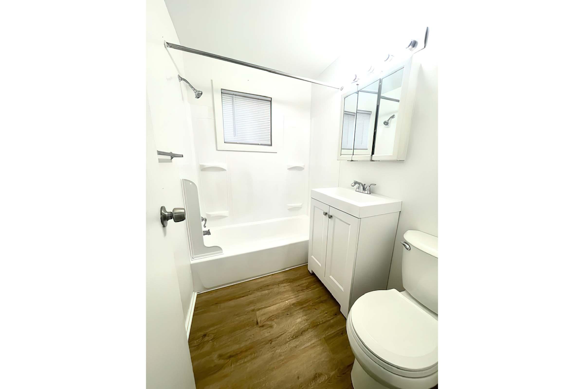 A clean, modern bathroom featuring a white bathtub with a shower curtain, a small white vanity with a sink, a toilet, and light-colored wood flooring. Natural light comes through a window above the tub, and there are bright lights above the mirror. The walls are painted a light color.