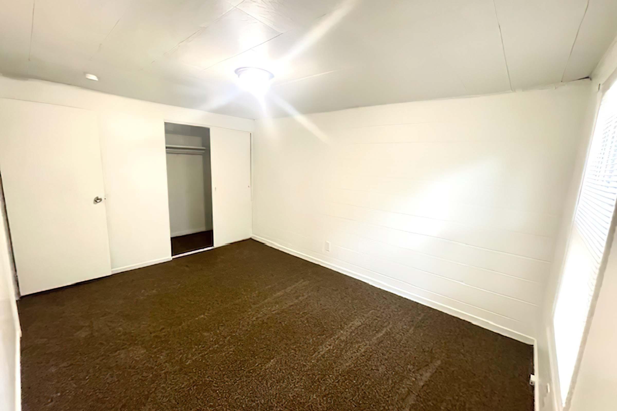 A small, empty room with light-colored walls and a dark brown carpet. There is a single light fixture on the ceiling and a closet with sliding doors. A window is visible on the right side, allowing natural light into the space. The room is clean and uncluttered, creating a minimalistic atmosphere.