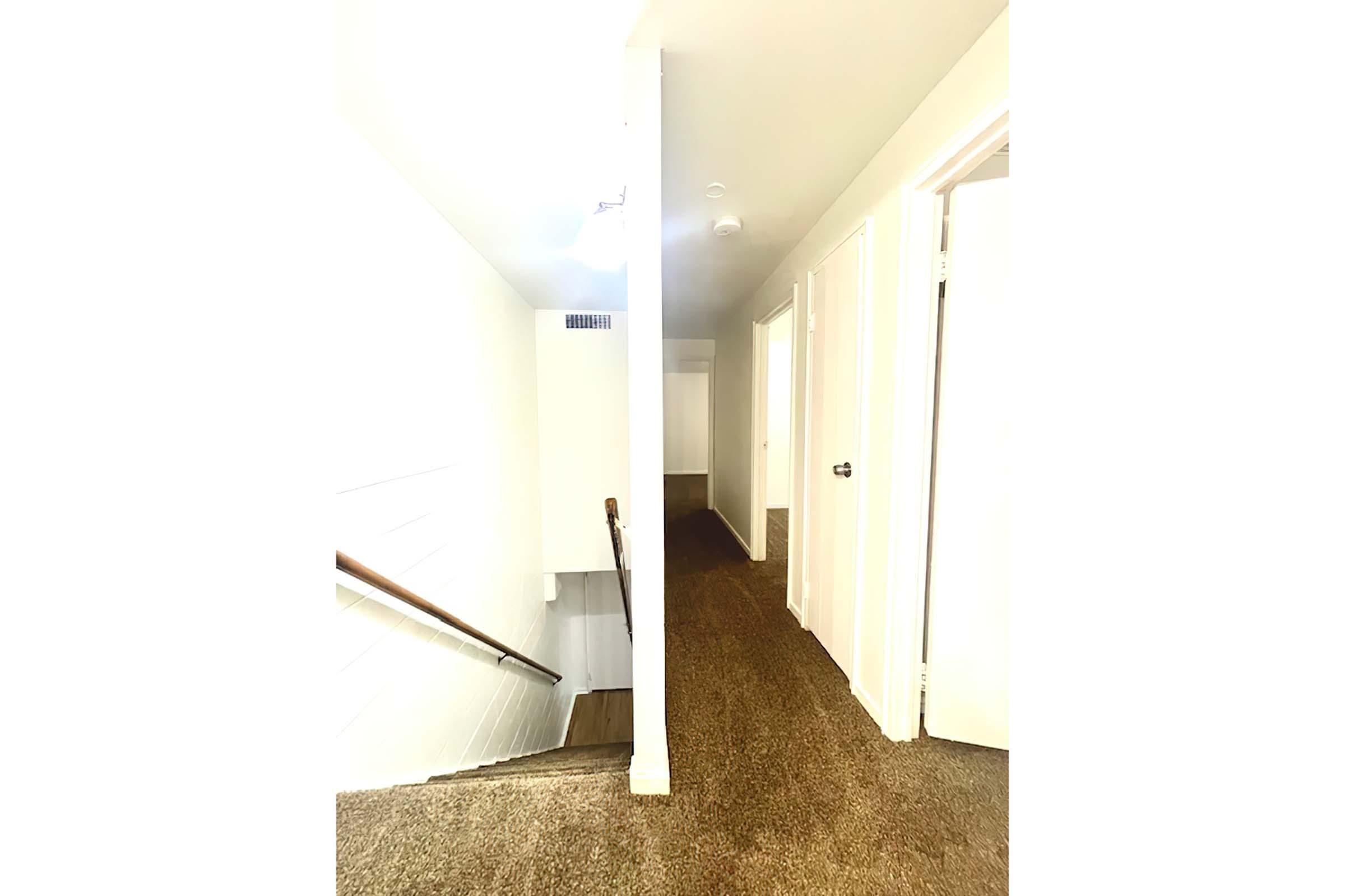 A well-lit hallway with carpeted flooring, leading to a staircase on the left. The walls are painted white, and there are several closed doors along the right side. The space appears clean and modern, with natural light coming from the top of the stairs.