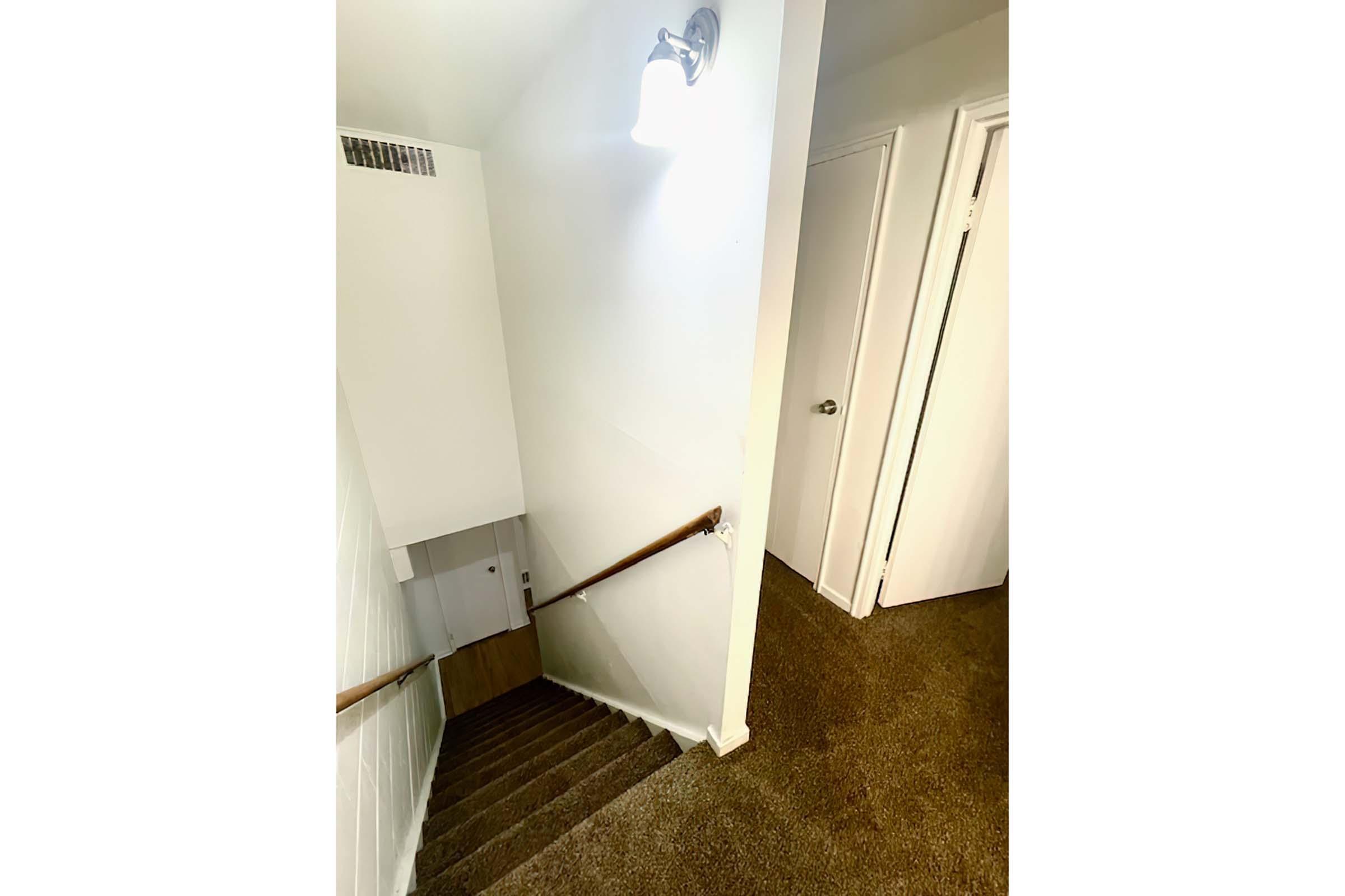 A well-lit staircase leading down with a wooden handrail, featuring carpeted steps. At the bottom of the stairs, there is an open area with a white wall and a door on the right. The light fixture above provides illumination, enhancing the inviting atmosphere of the space.