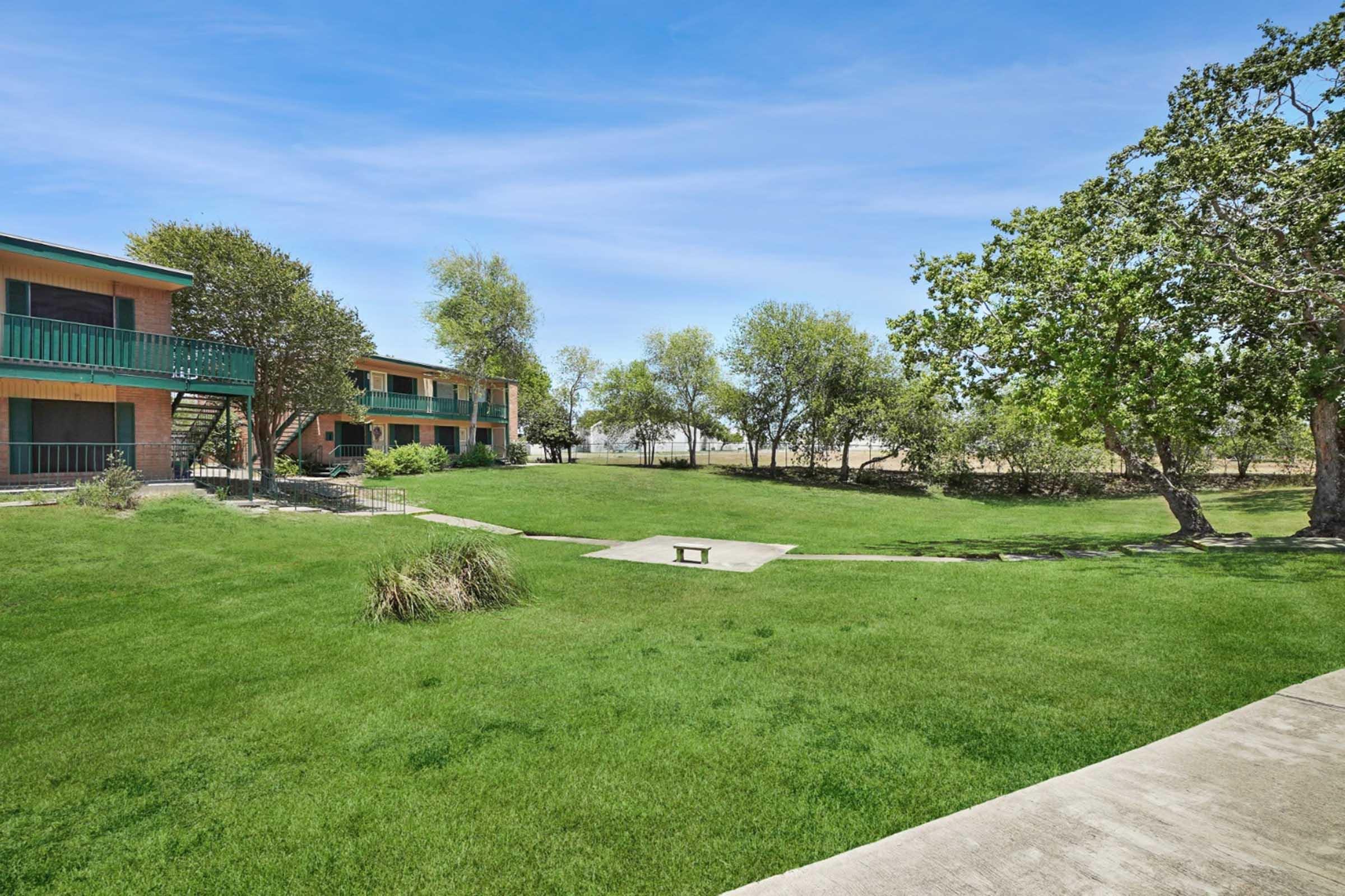 A green lawn area with sparse trees and a concrete path, leading to a two-story building with balconies. The building features a brick exterior and blue railings. The scene is set under a clear blue sky, suggesting a tranquil outdoor environment.