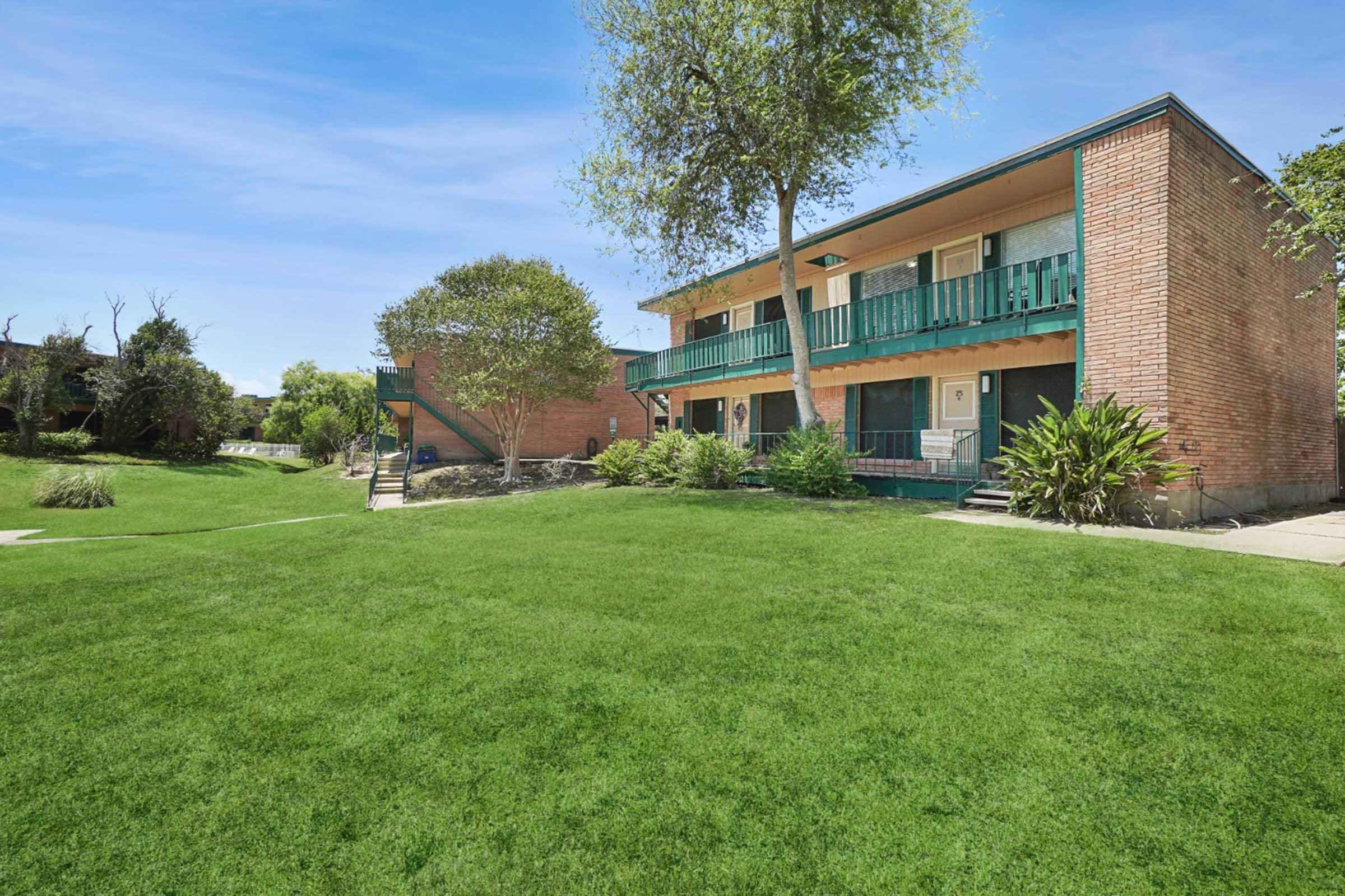 Two-story brick apartments with green balconies set in a landscaped area featuring lush grass and trees. A pathway leads to the entrance, and there are plants surrounding the building, creating a peaceful and inviting atmosphere.