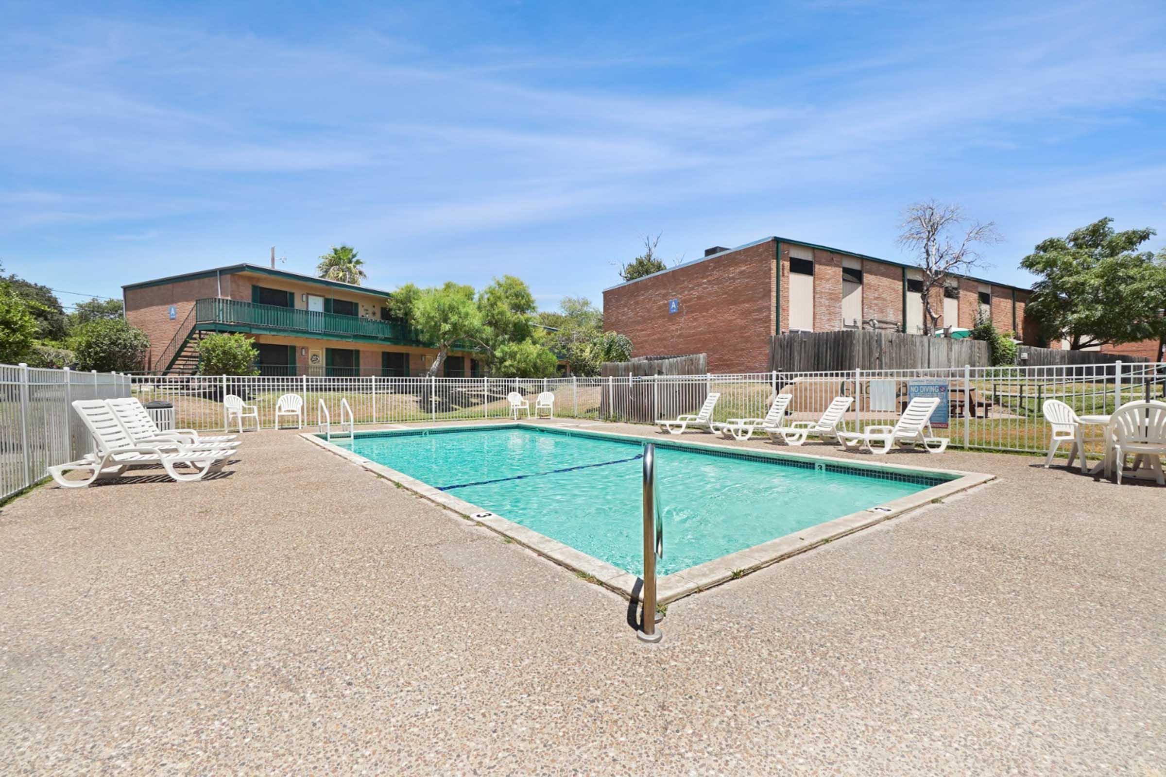 A clear swimming pool surrounded by a concrete deck, with several white lounge chairs positioned around it. In the background, there are two multi-story brick apartment buildings and greenery, under a bright blue sky.