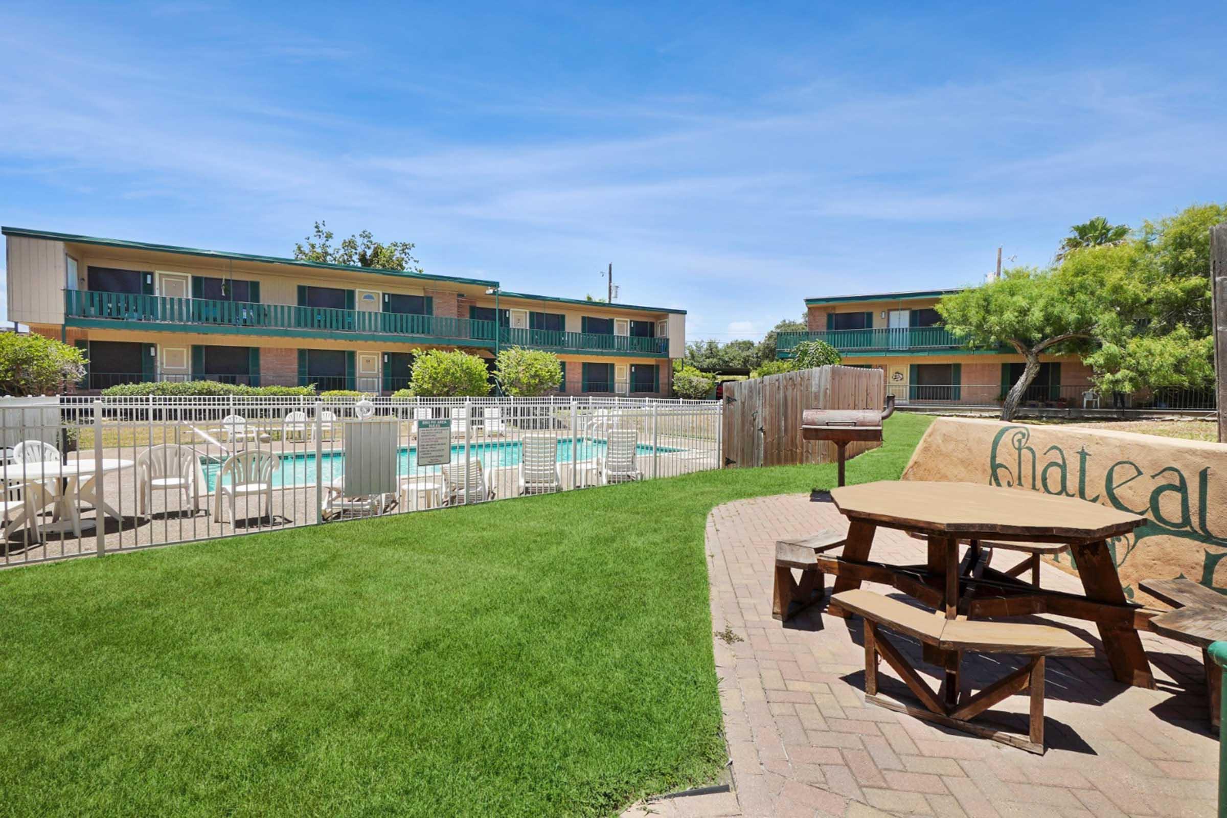 View of a motel featuring a swimming pool surrounded by lounge chairs. In the foreground, a picnic area with wooden tables is set on green grass, with the motel buildings visible in the background, showcasing a serene outdoor environment. The sky is clear and blue, adding to the inviting atmosphere.