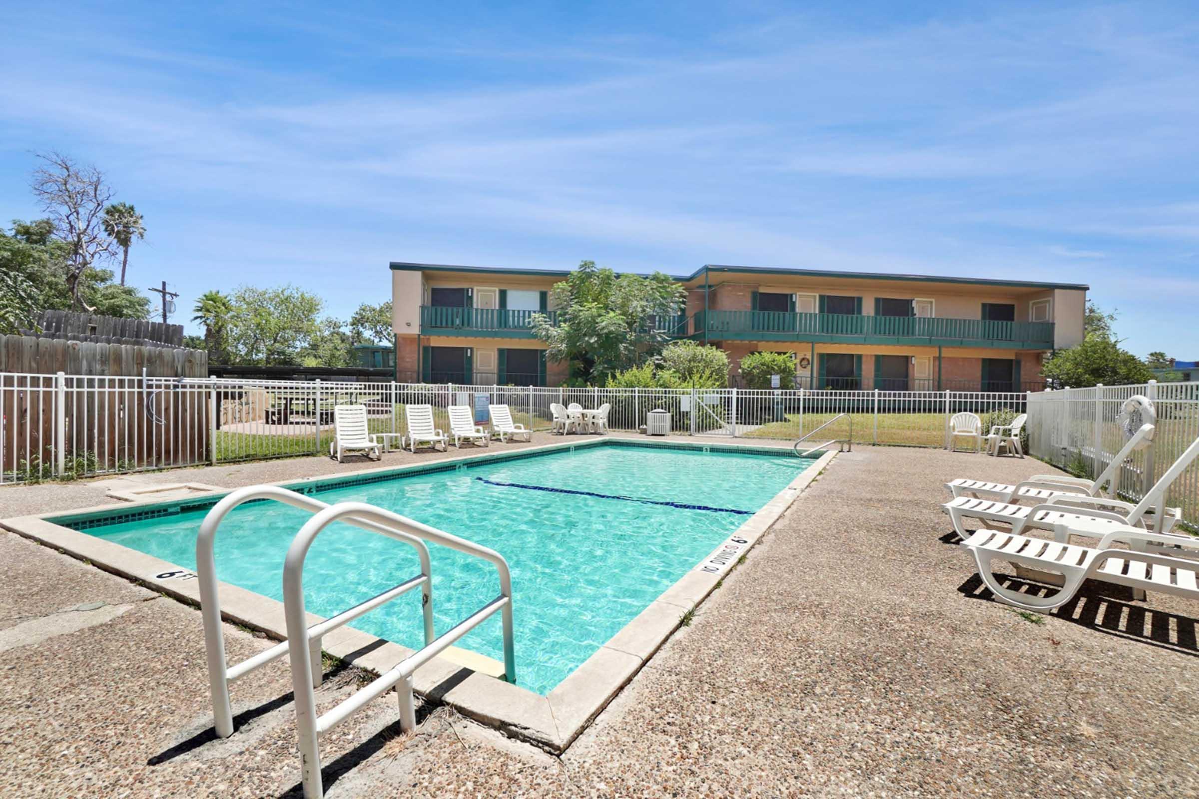 A clear swimming pool surrounded by lounge chairs, with a gated area in a sunny setting. In the background, a two-story building features green balconies and lush greenery. The scene conveys a relaxing atmosphere perfect for leisure and enjoyment.