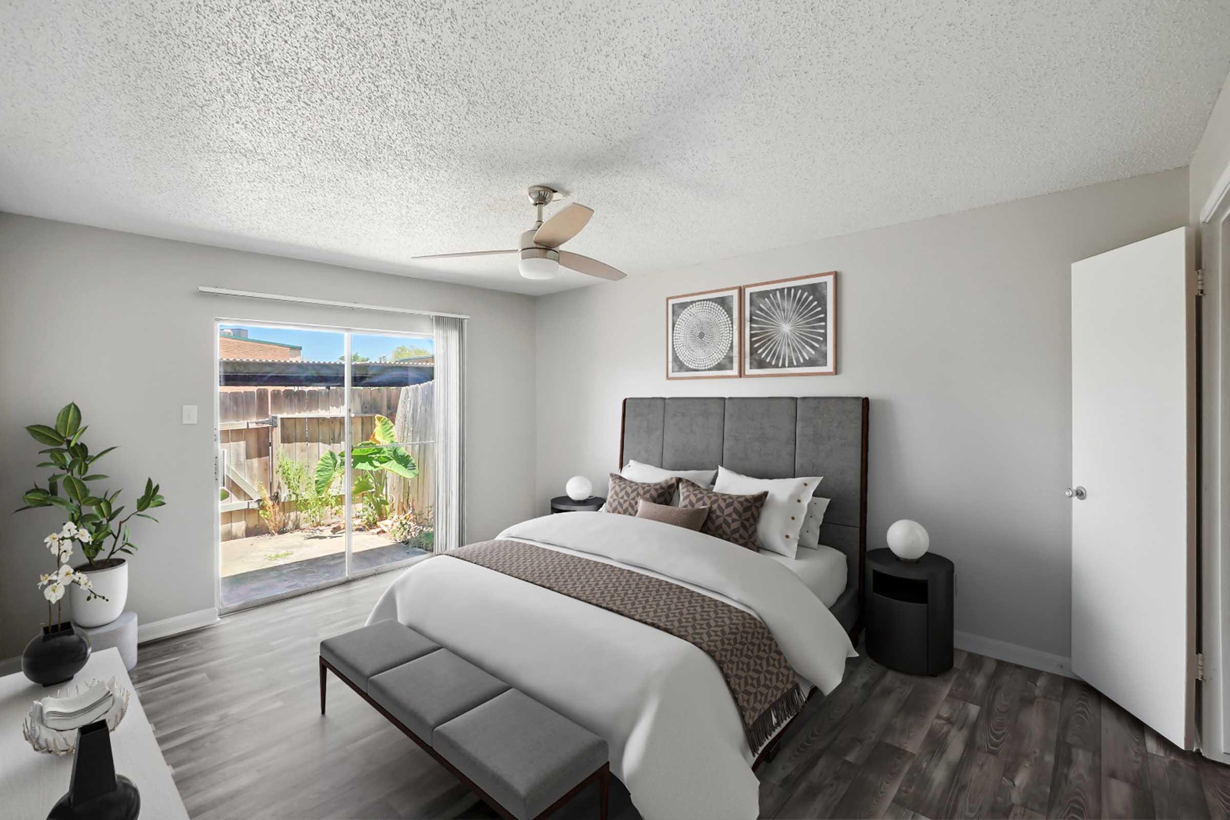 A well-lit, modern bedroom featuring a grey bed with decorative pillows and a quilt. There's a bench at the foot of the bed, a plant in the corner, and framed artwork on the wall. A sliding glass door leads to an outdoor area, enhancing the room's airy feel.