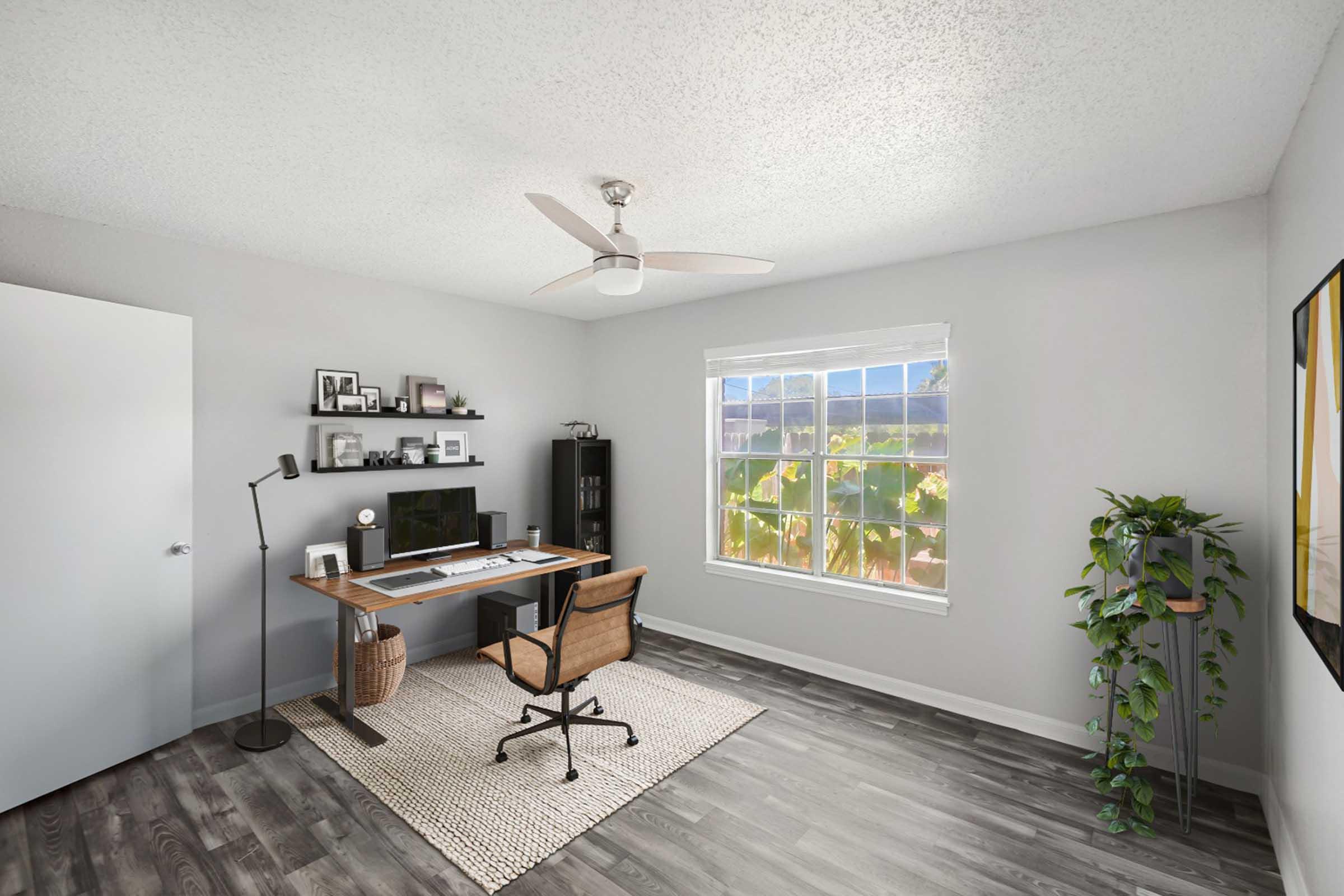 A modern home office featuring a desk with a computer, a comfortable chair, and a lamp. A wall shelf holds framed photos and a decorative plant. A window allows natural light to fill the room, showcasing greenery outside. The space is painted in light colors with wooden flooring and a cozy area rug.