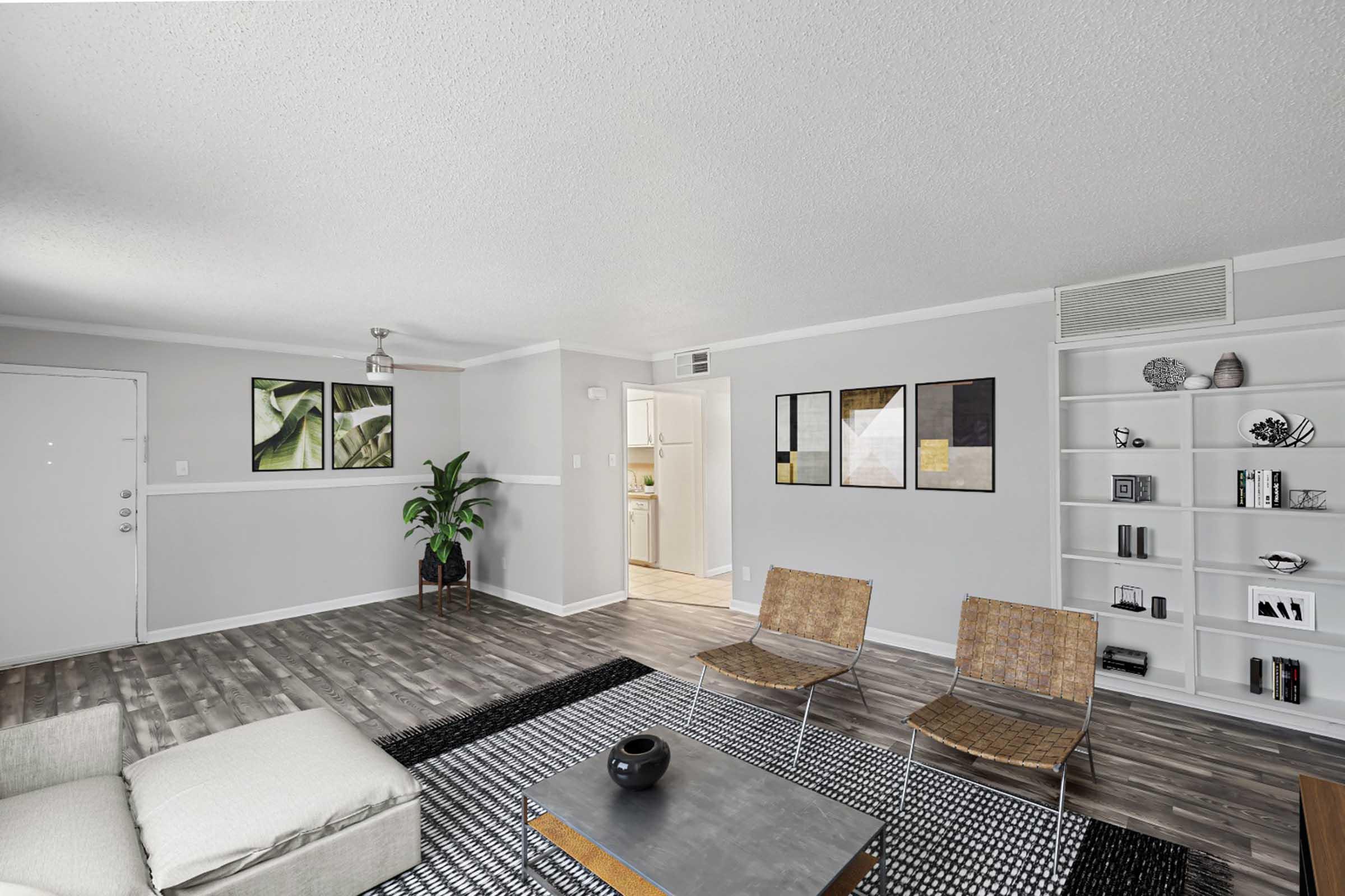 A modern living room featuring a gray couch, two woven chairs, and a coffee table with a black decorative bowl. The walls are painted light gray with framed artwork, while a plant adds a touch of greenery. A bookshelf displays decorative items, and a doorway leads to a kitchen area in the background.