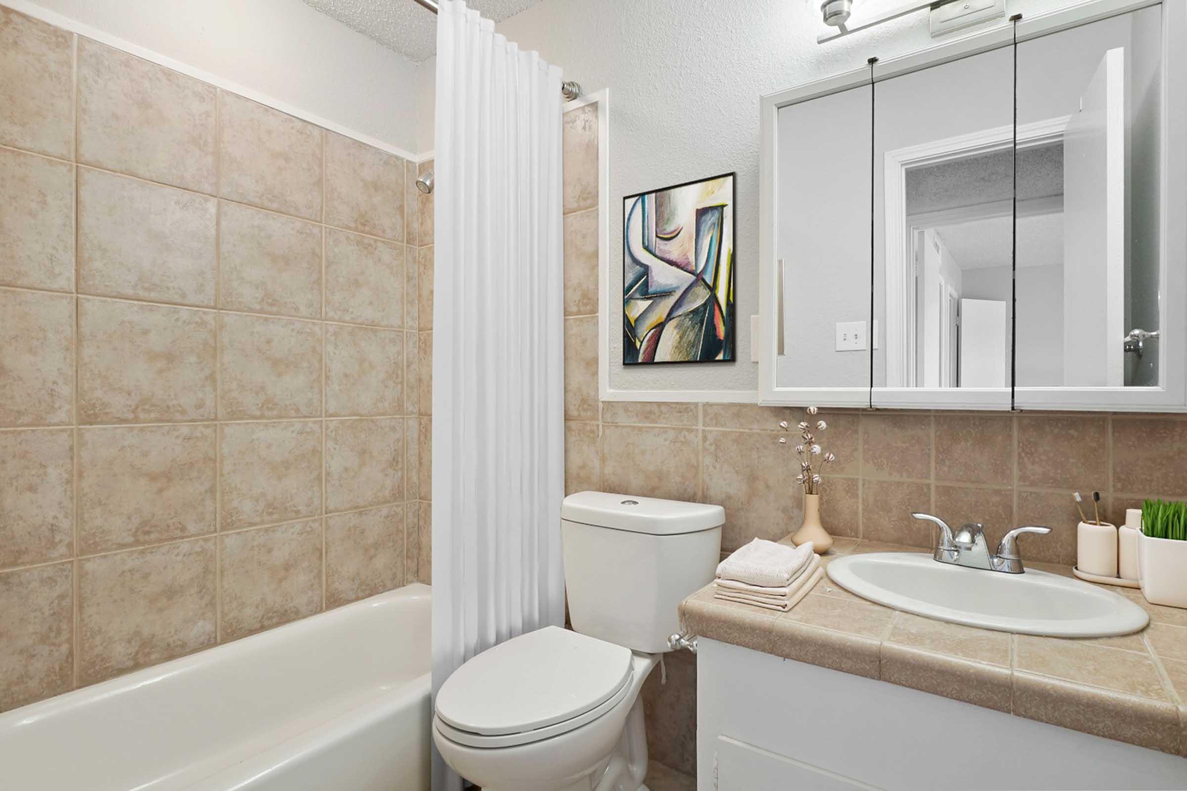 A clean and modern bathroom featuring a bathtub with a shower, a white toilet, and a round sink with a mirror above. The walls are tiled in beige, and there is an abstract art piece on the wall. A small vase with flowers and neatly arranged towels add a touch of decor.