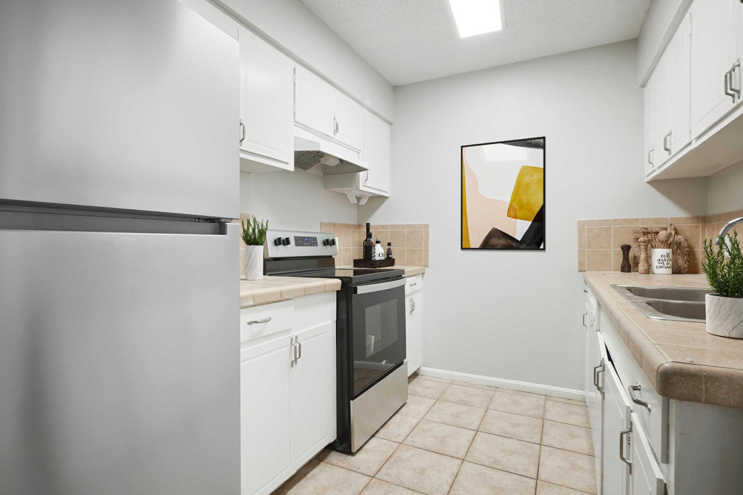A modern kitchen featuring white cabinetry, stainless steel appliances, and beige tile flooring. An oven and stove are positioned between a refrigerator and a sink. The walls are light gray, and a colorful abstract painting hangs on one wall, adding a touch of decor. Potted plants are on the countertops.