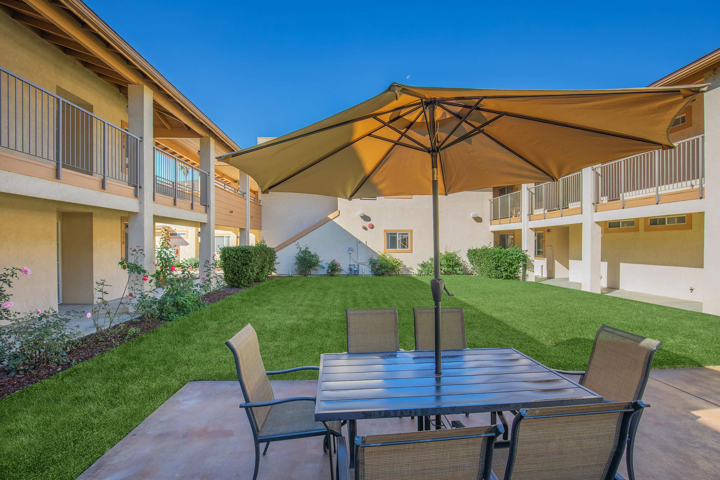 A sunny courtyard featuring a large umbrella-shaded table and chairs. Surrounding the area are well-maintained grass, flower beds, and two-story buildings with balconies. The clear blue sky complements the inviting outdoor setting, ideal for relaxation or social gatherings.