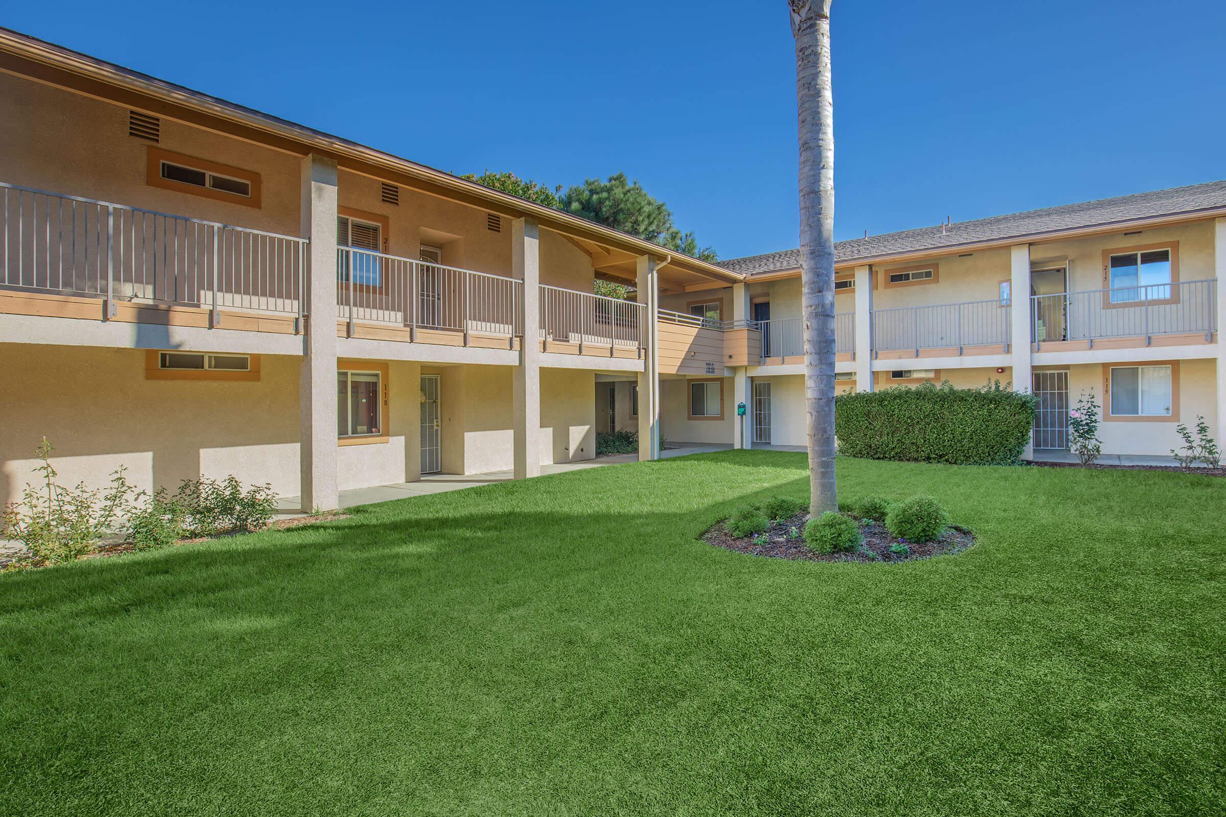 A view of a courtyard in a residential complex featuring two-story buildings with balconies, surrounded by green lawns and small bushes. A palm tree stands in the center, and the sky is clear and blue, creating a bright and inviting atmosphere.