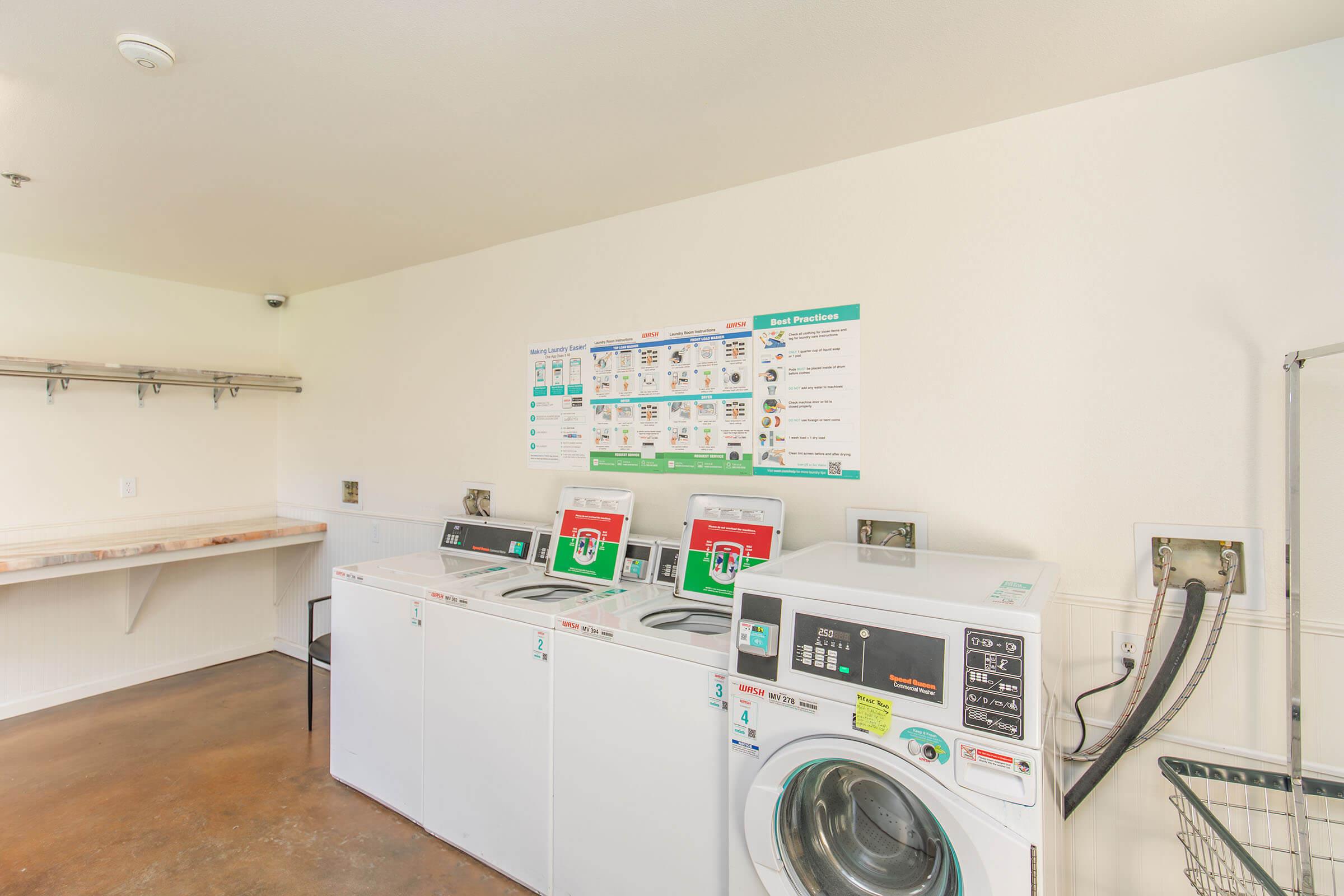 A clean, bright laundry room featuring multiple washing machines and a dryer. Instructions and guidelines are posted on the wall, and there is a folding counter and hooks for hanging clothes. The flooring is a polished concrete.
