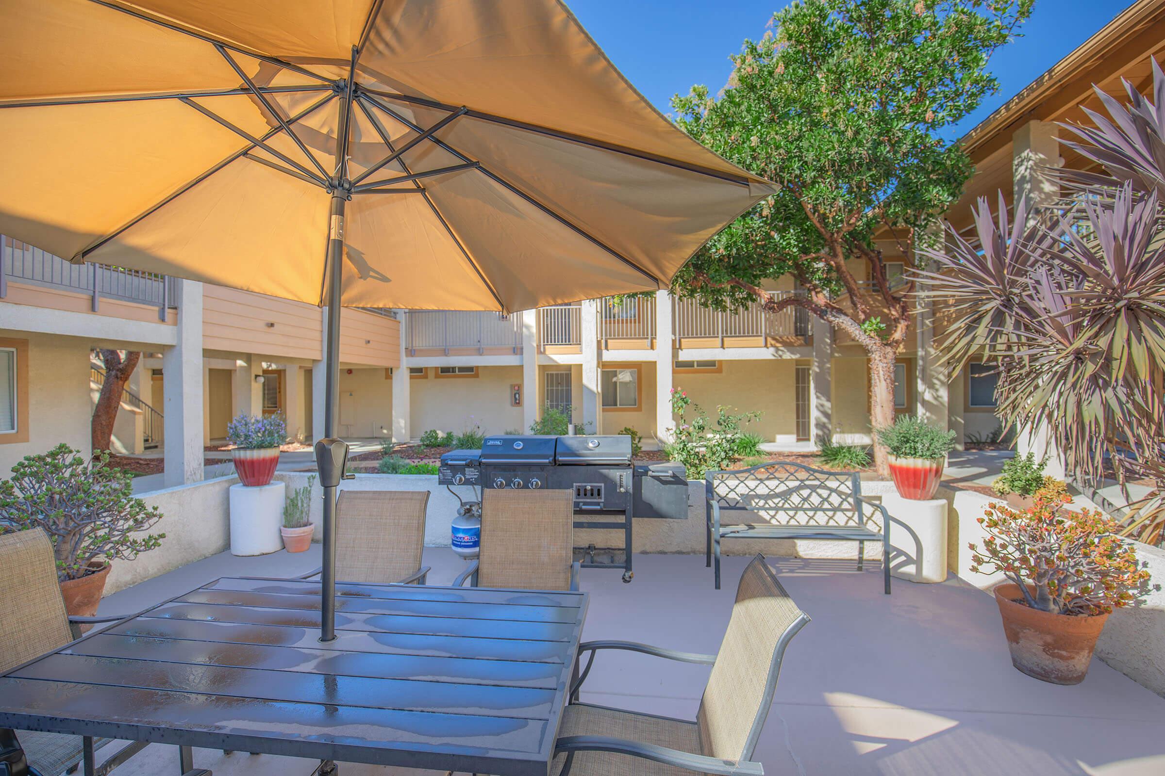 A sunlit outdoor patio featuring a large umbrella over a black dining table and chairs. In the background, there is a barbecue grill and various potted plants. Surrounding buildings are visible, adding to the cozy atmosphere of the space.