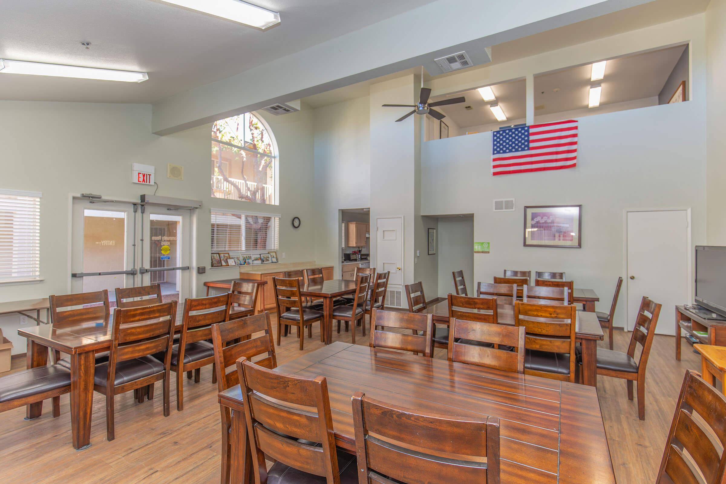 A spacious dining area featuring multiple wooden tables and chairs, with large windows allowing natural light. An American flag hangs on the wall, and there is a ceiling fan for ventilation. The room has a warm ambiance, suitable for gatherings or meals.