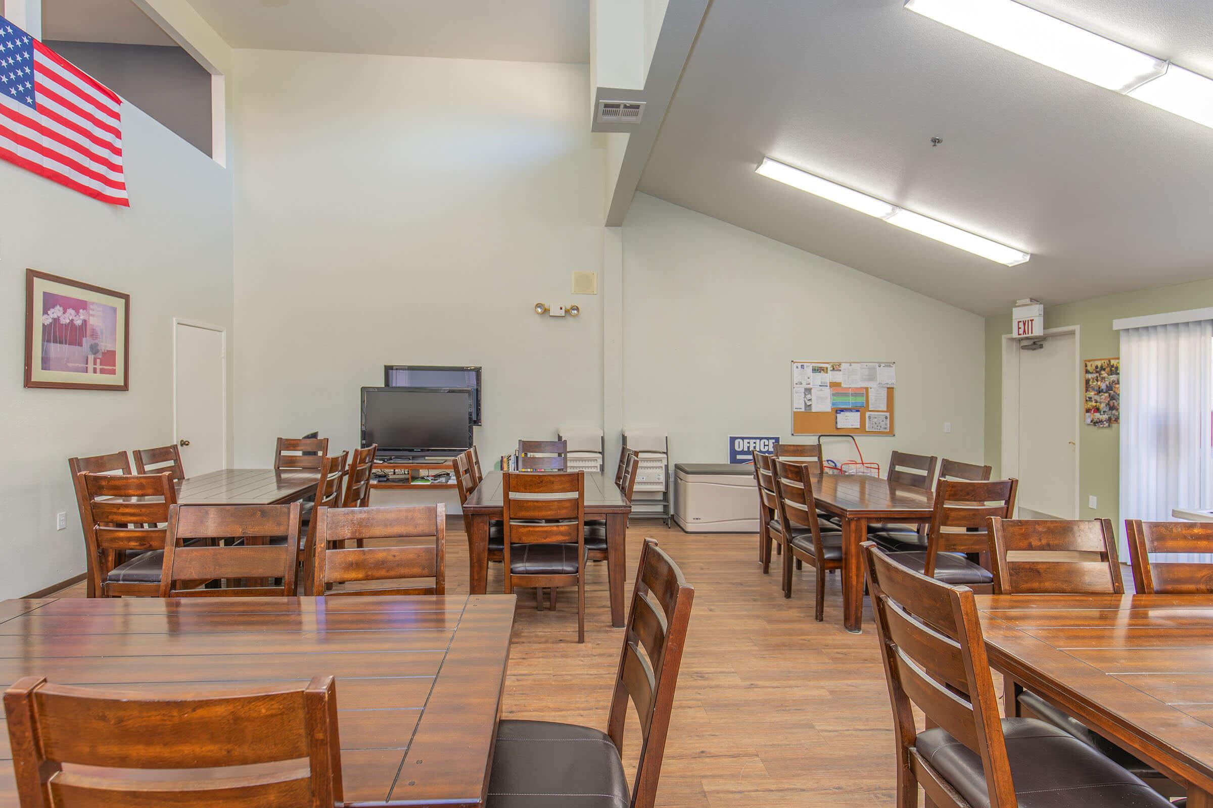 Interior view of a room featuring wooden dining tables and chairs. There is a television on a stand, a bulletin board on the wall, and an exit sign visible. A large window allows natural light to fill the space. An American flag is displayed on the wall, contributing to a communal atmosphere.