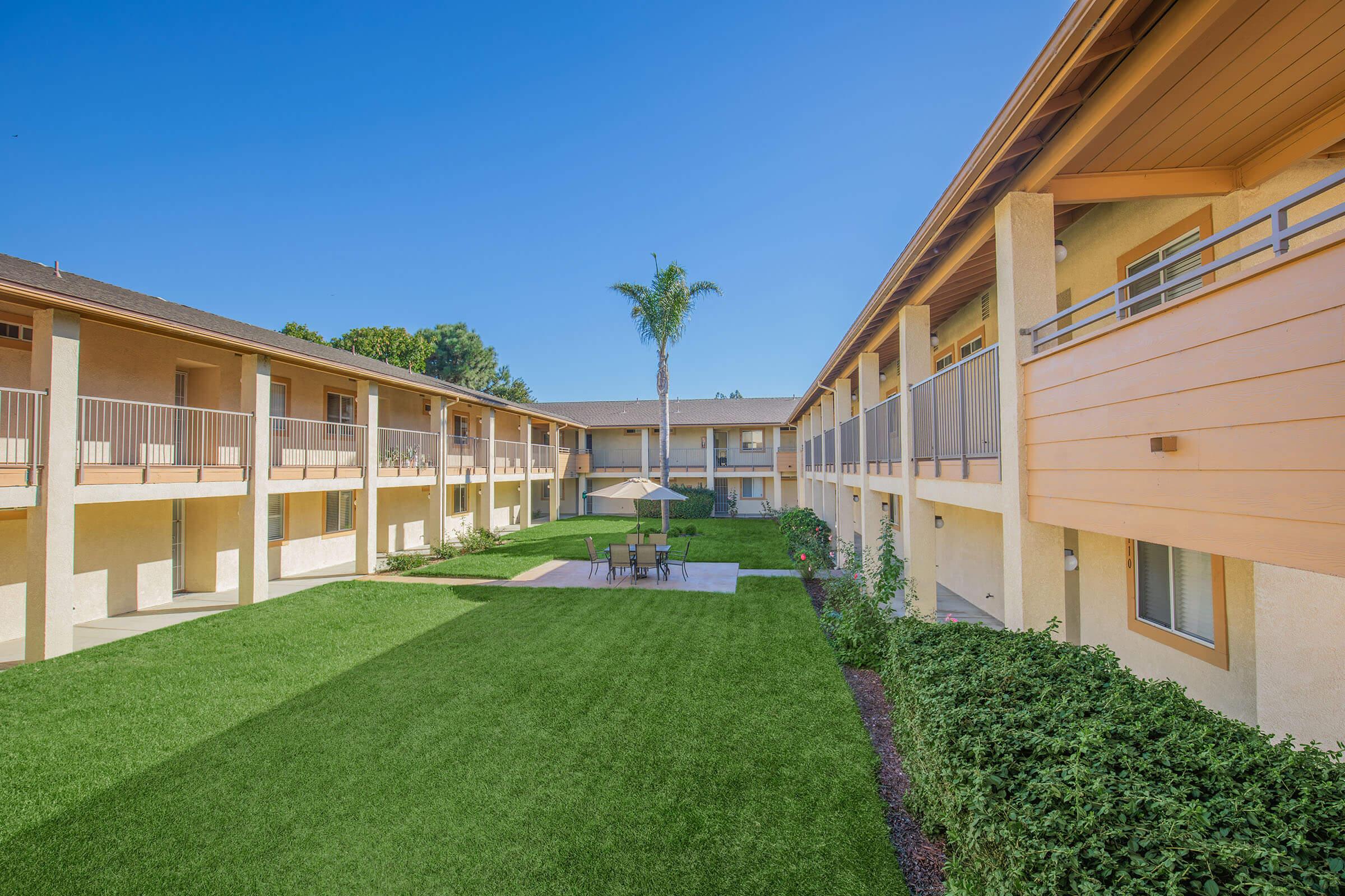 A view of a well-maintained courtyard in an apartment complex. The area features a grassy lawn with a table and chairs under an umbrella, surrounded by two-level buildings with balconies. Lush green shrubs line the pathways, and a palm tree stands prominently, set against a clear blue sky.