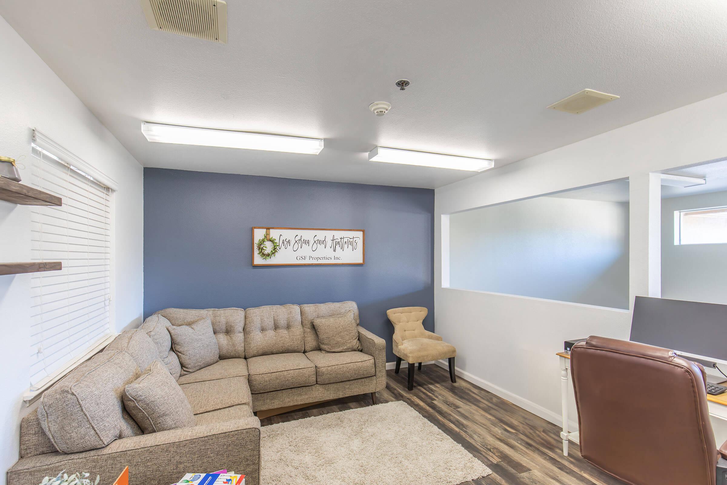 A cozy office space featuring a brown sectional sofa, a beige chair, and a light-colored rug. Walls are painted in a combination of white and dark blue. There's a wall-mounted sign that reads, "New Year, New Beginnings." Natural light comes through a window, creating a warm and inviting atmosphere.