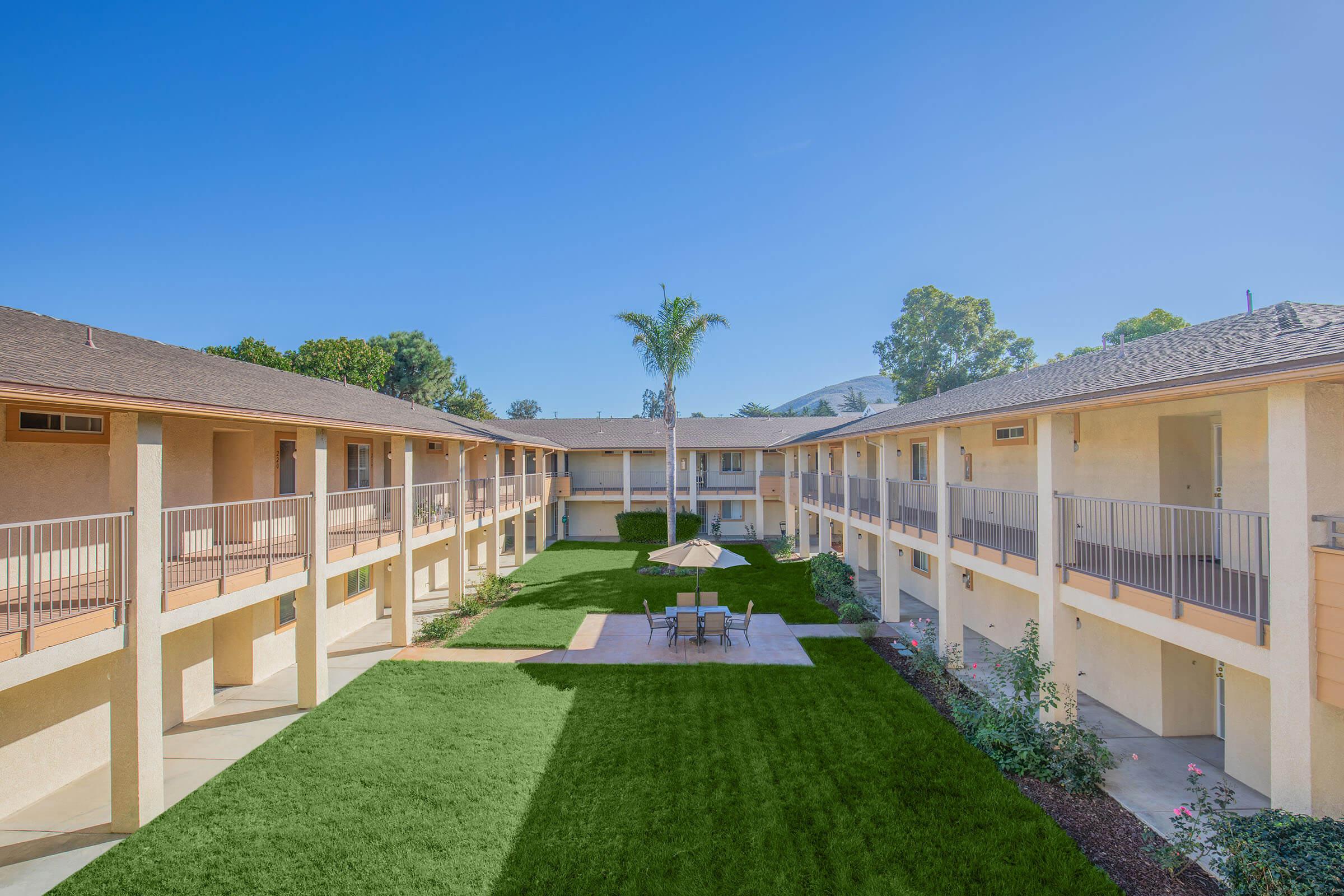 View of a multi-unit residential complex featuring two-story buildings arranged around a green courtyard. The courtyard includes a small table and chairs under an umbrella, surrounded by well-maintained grass and shrubbery. The sky is clear with bright sunlight illuminating the space.
