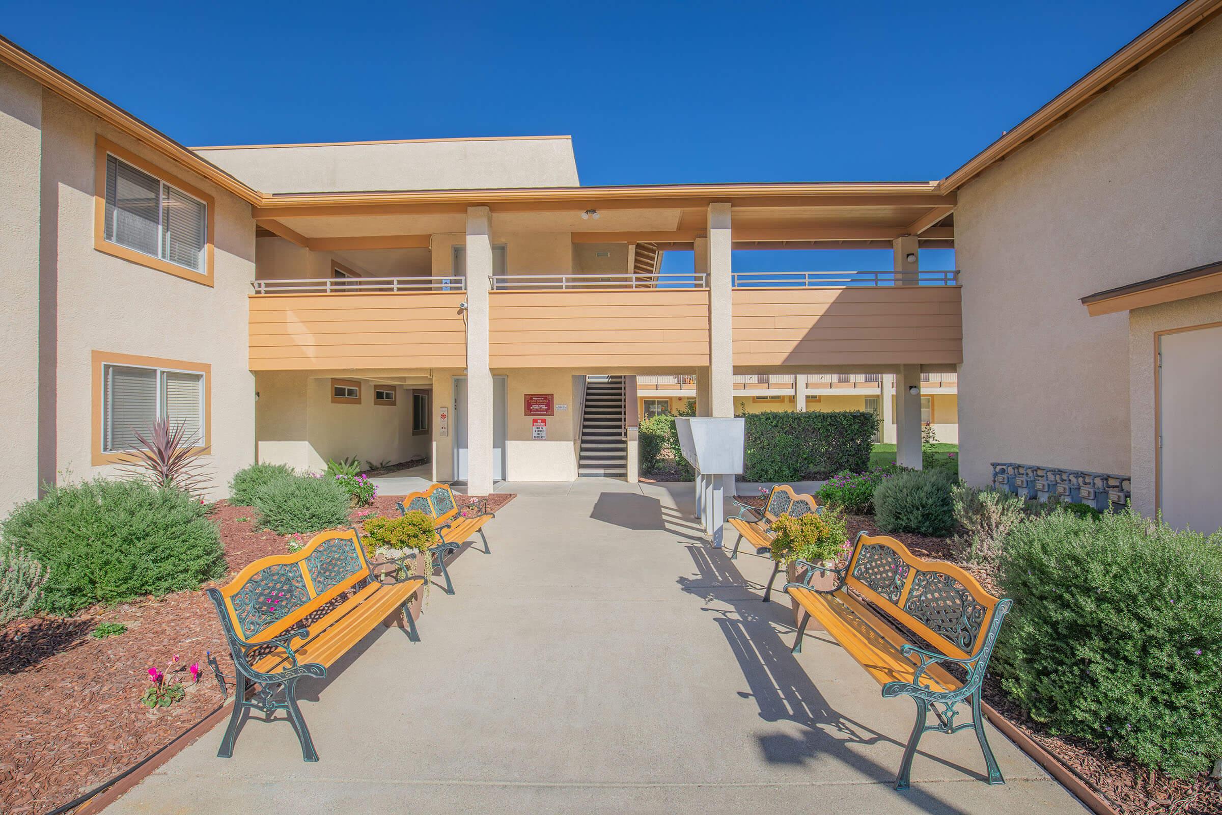 A well-lit walkway between two buildings featuring benches and landscaped shrubs. The entrance includes a staircase leading to an upper level, and the clear blue sky overhead enhances the bright, welcoming atmosphere of the area.