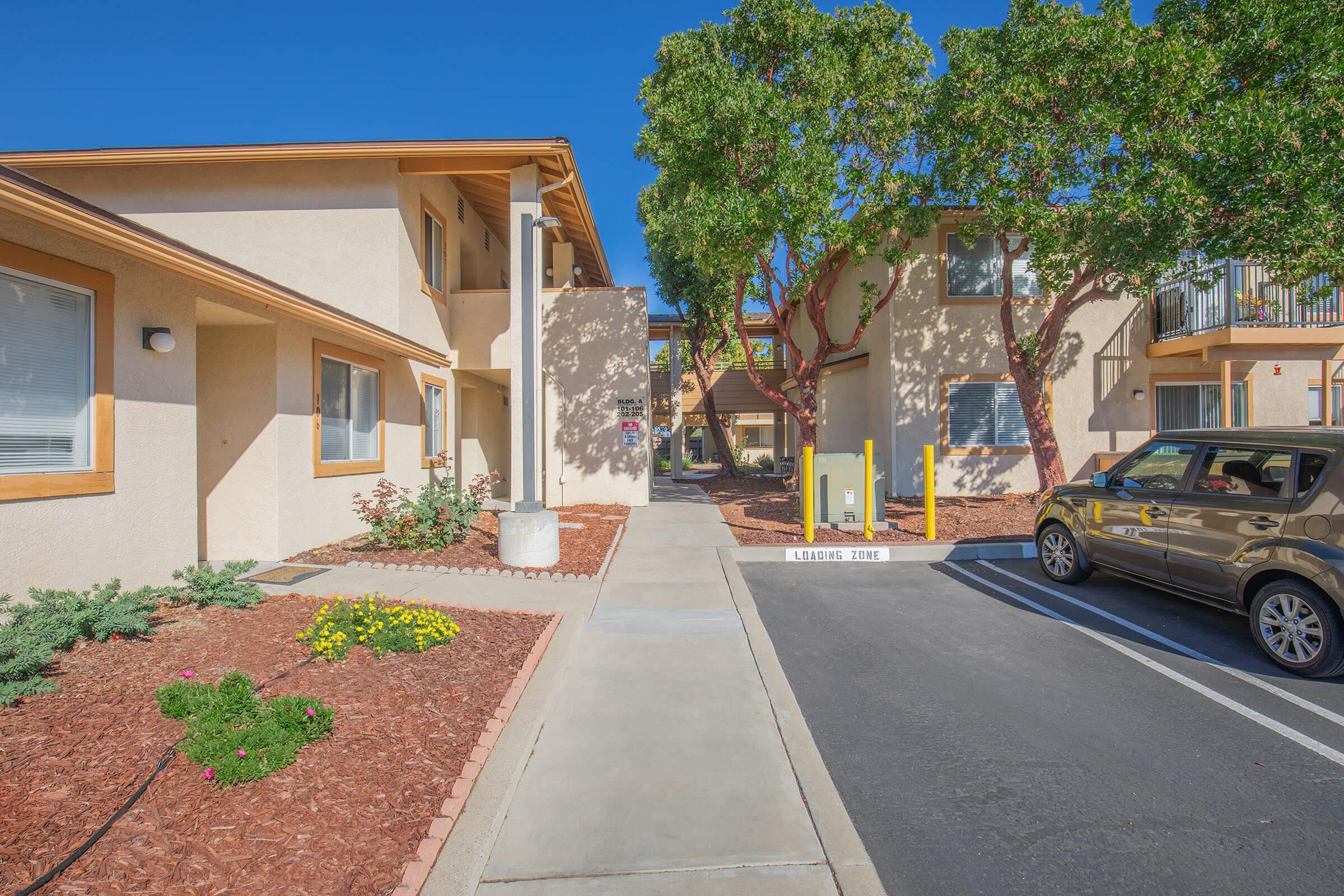 View of a residential complex with two-story buildings. The pathway leads between the structures, flanked by landscaped areas featuring flowers and shrubs. There are parking spaces on one side and trees providing shade, under a clear blue sky.