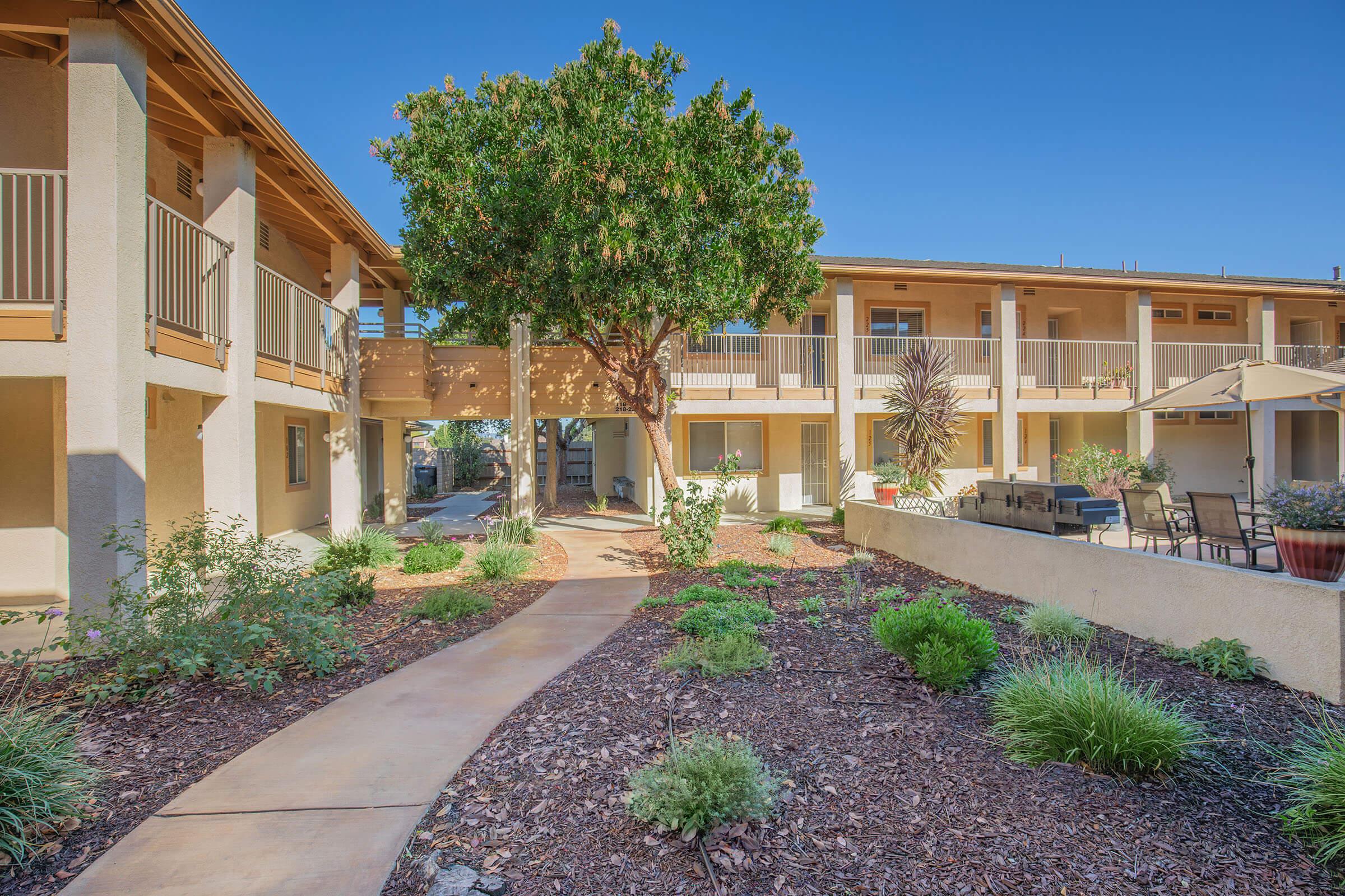 A peaceful outdoor area featuring a landscaped garden with various plants and shrubs, a curved pathway leading through the space, and a few buildings with balconies. Sunlight illuminates the scene against a clear blue sky, creating a welcoming atmosphere. Outdoor seating is visible, inviting relaxation.