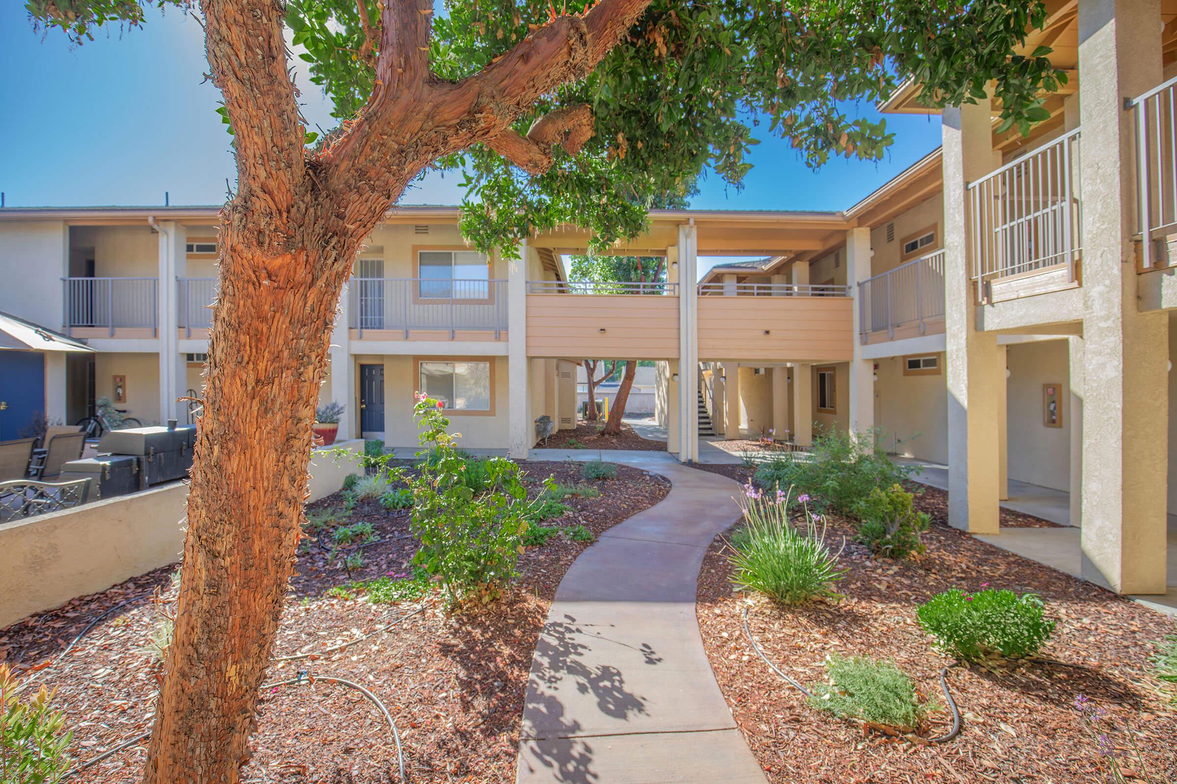 A landscaped courtyard featuring a winding path surrounded by greenery and plants, leading to a two-story building with multiple balconies. A tree adds shade to the area, creating a serene atmosphere in a residential complex.