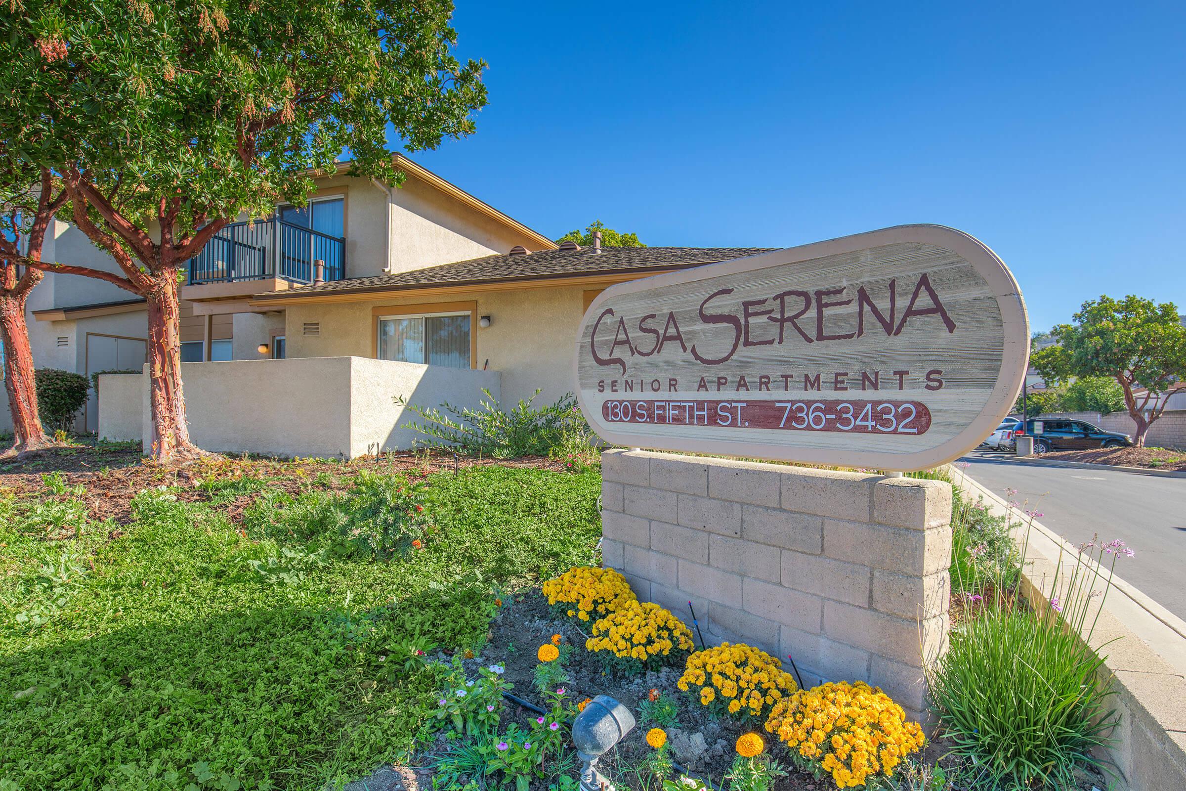 Sign for Casa Serena Senior Apartments, featuring the name prominently displayed. The building is set against a clear blue sky with greenery and colorful flowers in the foreground. A driveway leads to the entrance, enhancing the welcoming atmosphere of the residence.