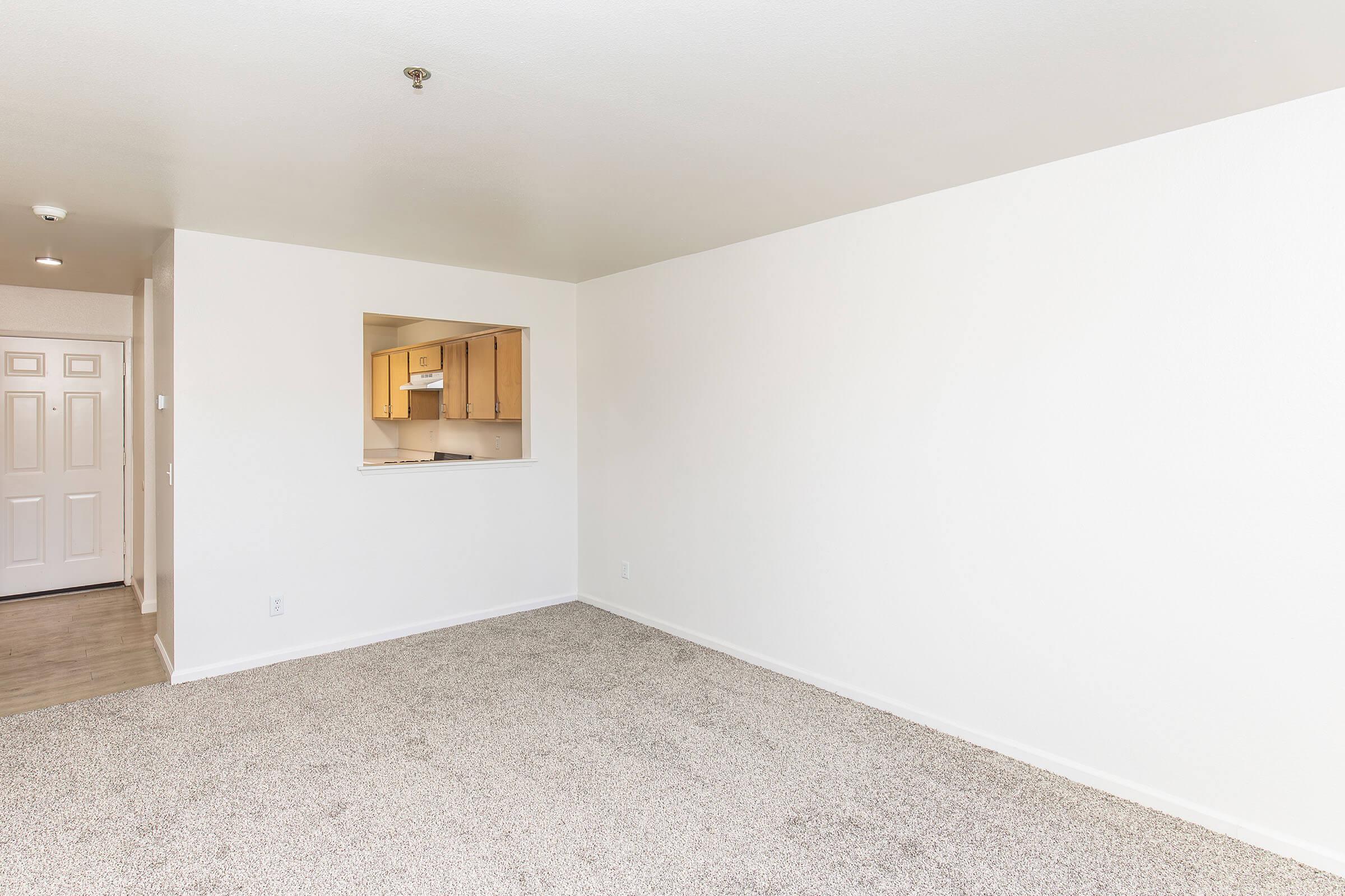 A clean, empty living space with light-colored walls and a textured carpet. There is a doorway on the left leading outside, and an open pass-through to a kitchen area in the background. The room is well-lit, creating a spacious and inviting atmosphere.