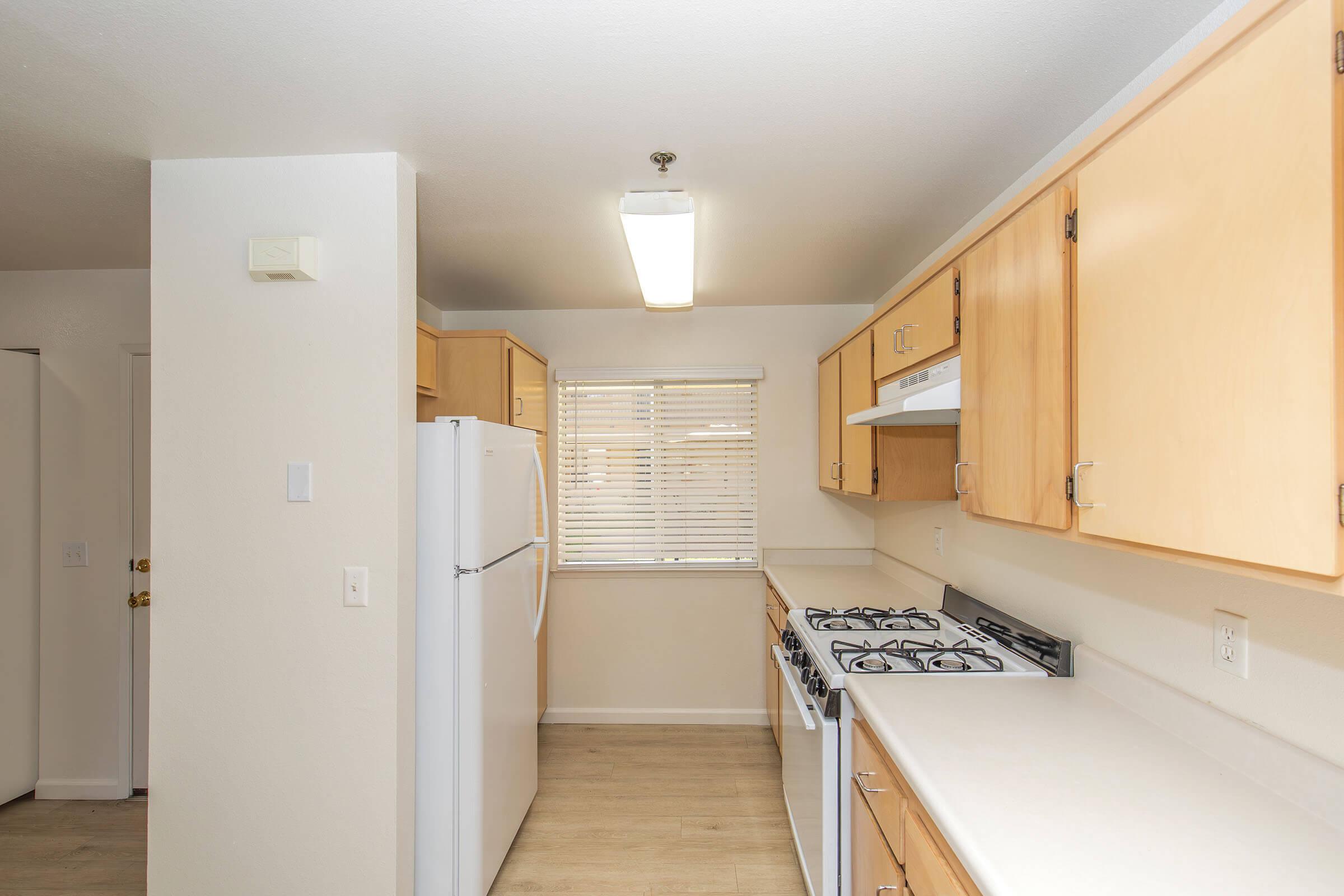 A bright, modern kitchen featuring light wooden cabinets, a white countertop, and a gas stove. A refrigerator is positioned on the left, and a window with blinds allows natural light to fill the space. The walls are painted in a neutral color, creating a warm and inviting atmosphere.