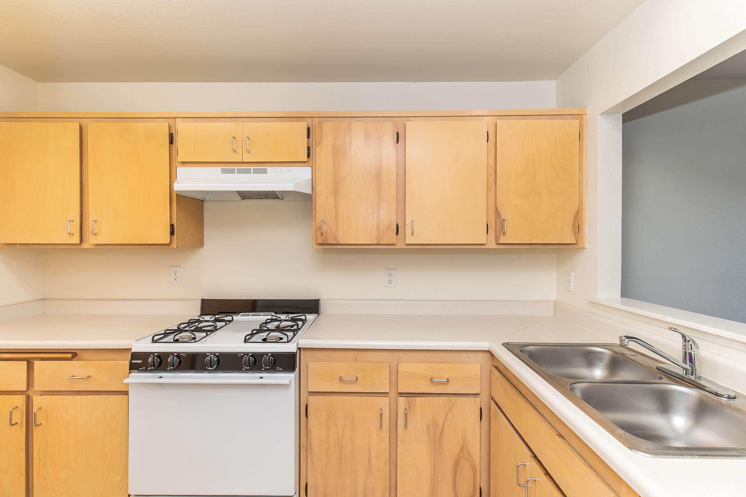 A clean, modern kitchen with light wooden cabinets and a white gas stove with five burners. There are dual stainless steel sinks near a window, providing natural light. The countertops are light-colored, creating a bright and inviting atmosphere.