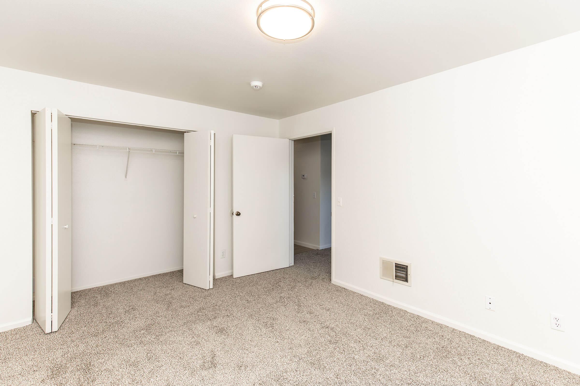 Empty room featuring light beige walls, carpeted flooring, and a ceiling light fixture. It includes two open doors leading to a closet and an adjoining room, providing a sense of spaciousness. The lack of furnishings emphasizes the room's potential for personal customization.