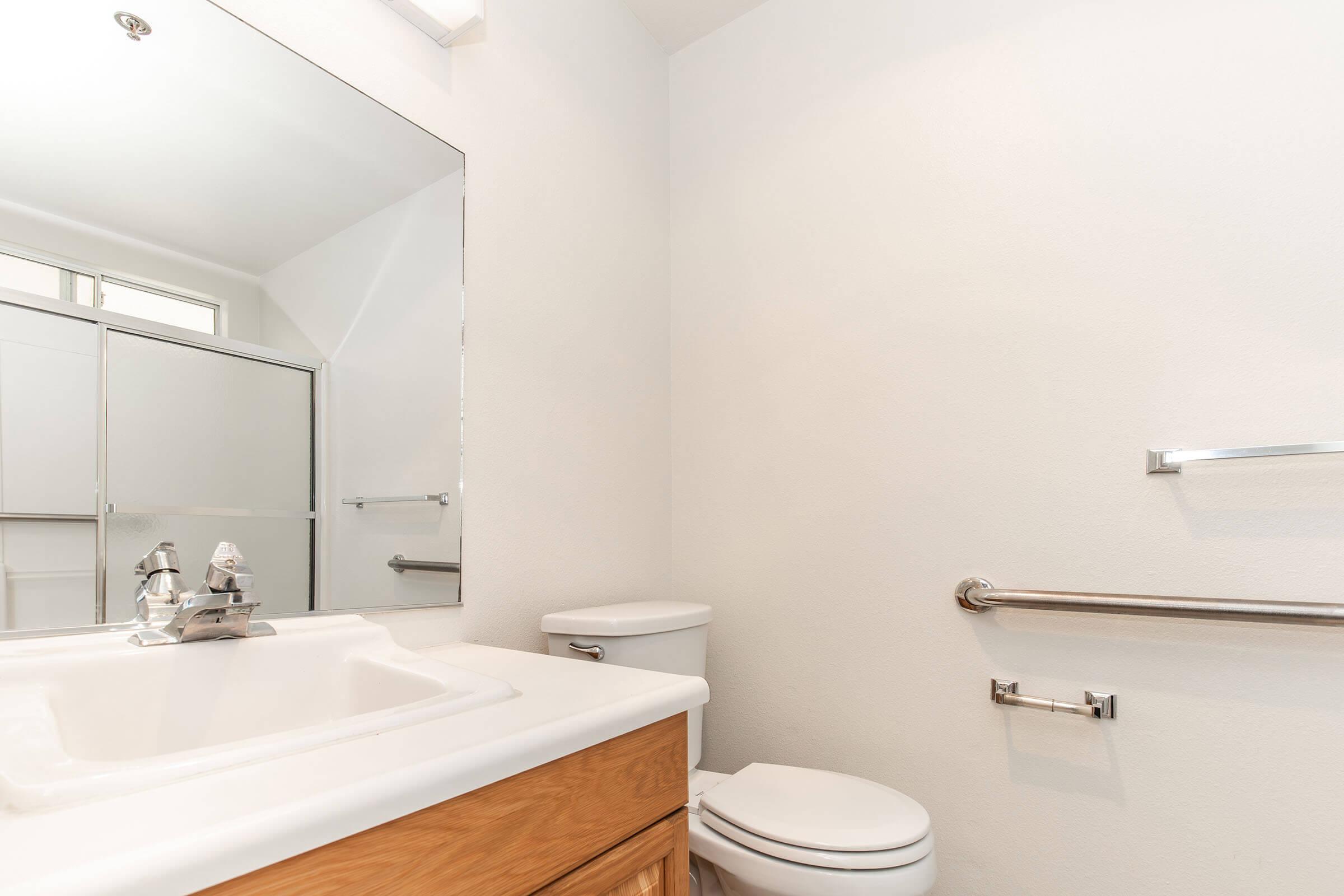 A clean, modern bathroom featuring a white countertop sink with a faucet, a large mirror, a toilet, and an accessible grab bar. The walls are painted in a light color, creating a bright and airy atmosphere. A clear shower enclosure is visible in the background.