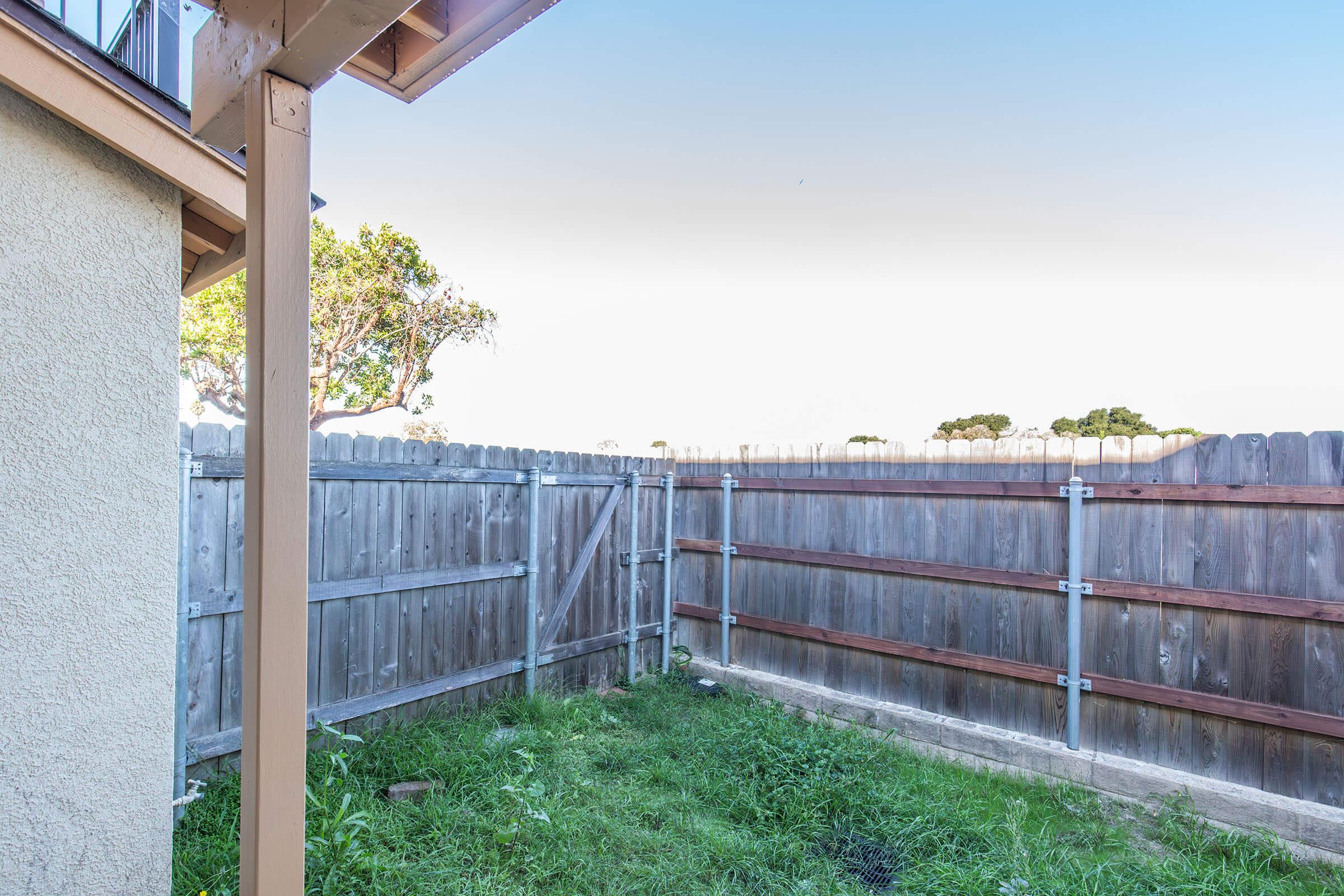 A fenced backyard area featuring a wooden fence with a gate, surrounded by grass. The view shows a clear sky and some trees in the distance, suggesting an open space. The structure of a house is visible in the corner of the image.