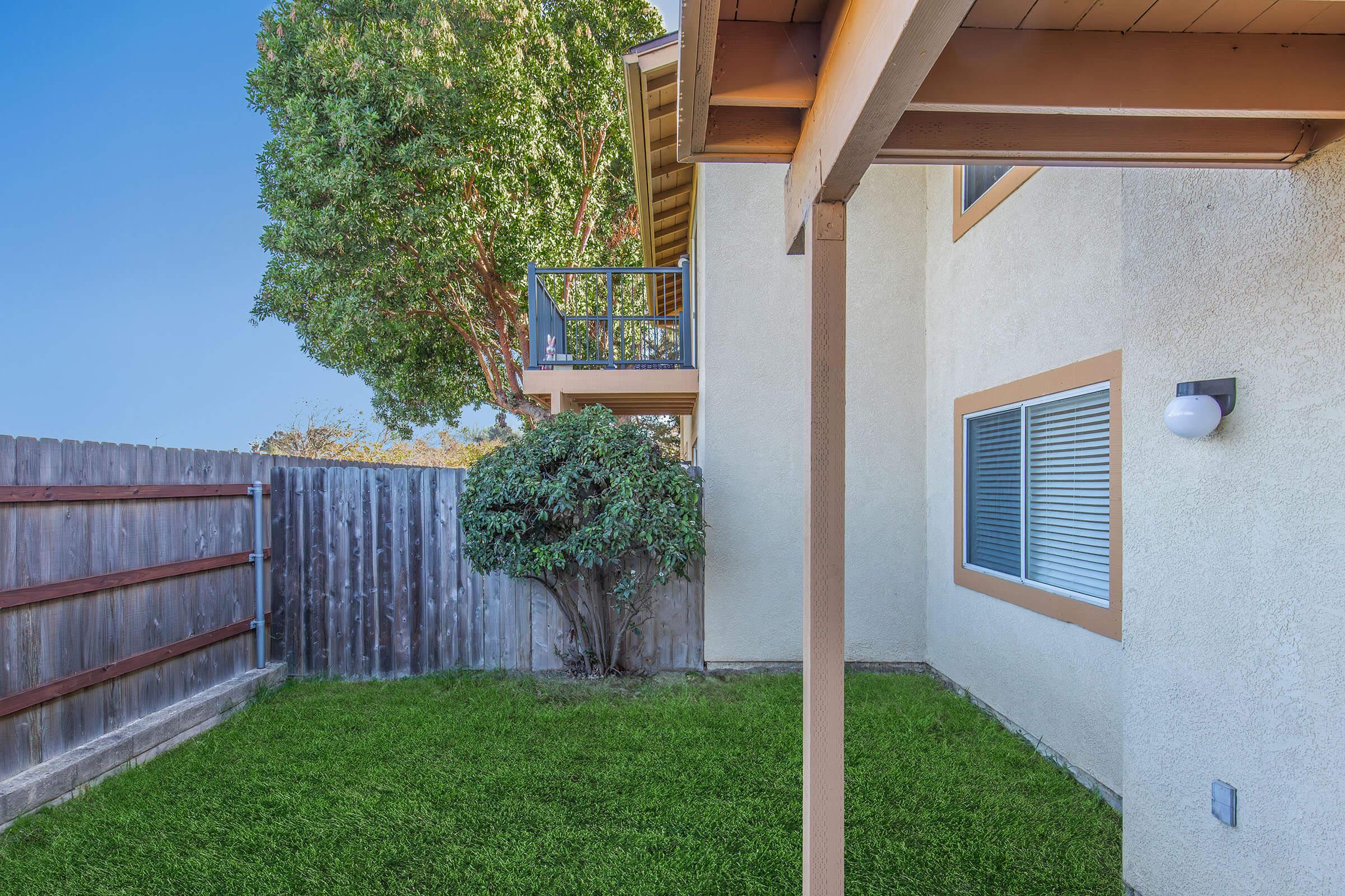 View of a side yard featuring a well-maintained green lawn, a small tree, and a wooden fence. Part of a neighboring building is visible, along with a balcony. Natural light illuminates the area, suggesting a pleasant outdoor space.