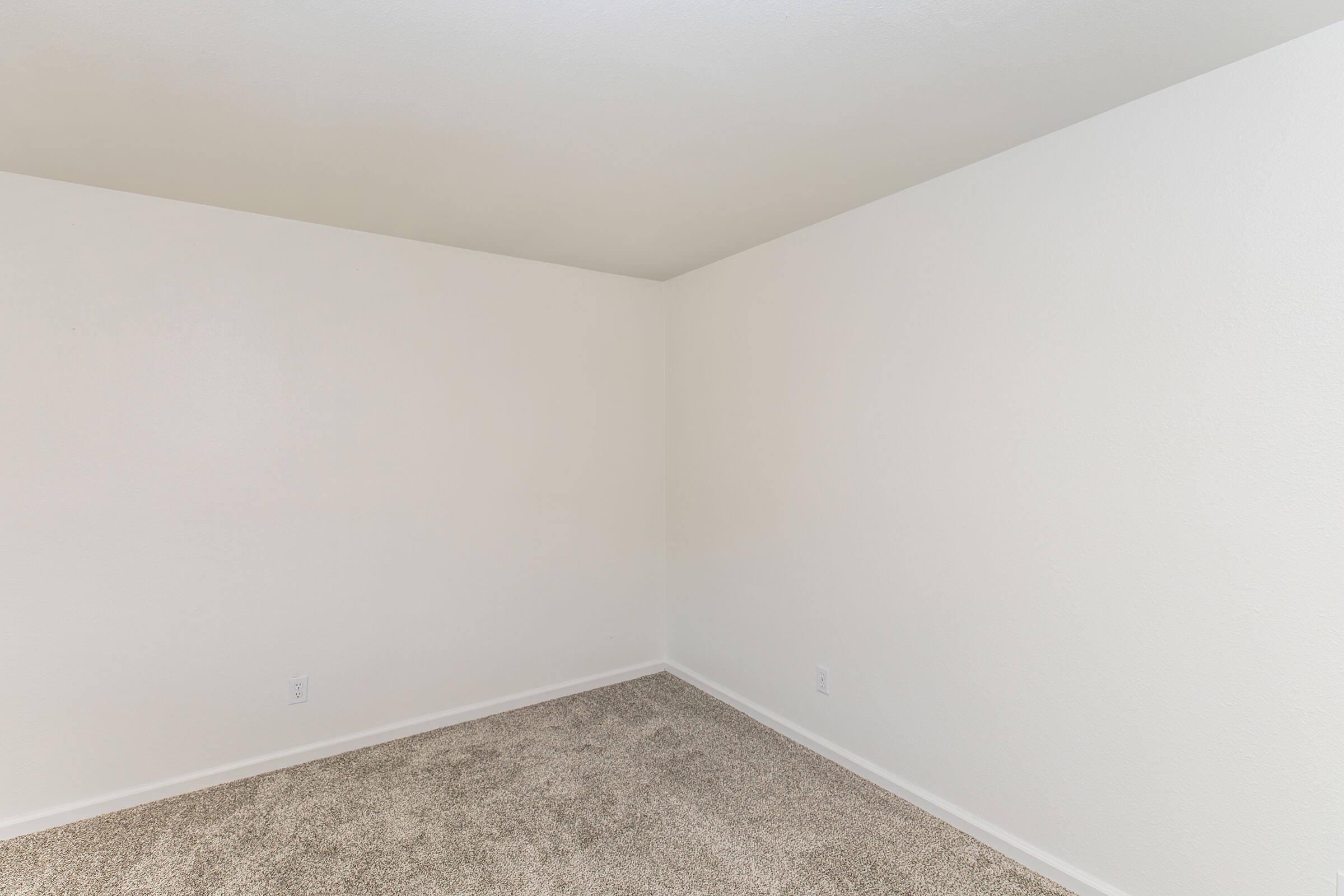 Empty room with light-colored walls and a carpeted floor. The room has a simple and minimalist design, featuring two bare walls and no furniture or decorations. The lighting is neutral, creating a bright and airy atmosphere.