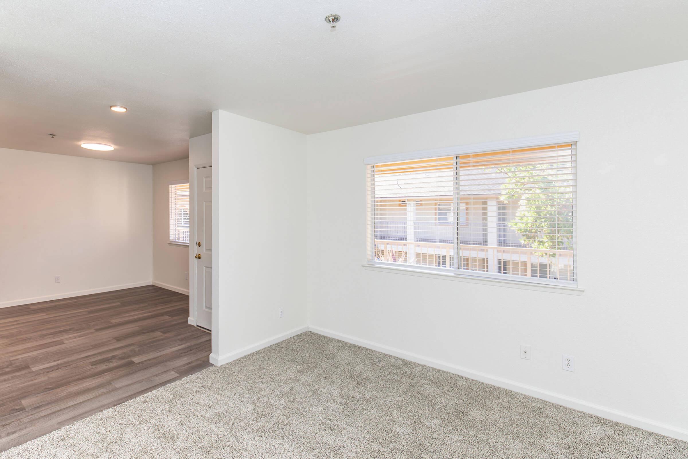 A spacious, empty room featuring light-colored walls and plush carpet. Large windows allow natural light to enter, enhancing the sense of openness. The adjoining area is visible, showcasing a similar clean and modern aesthetic with laminate flooring. Ideal for a living or dining space.