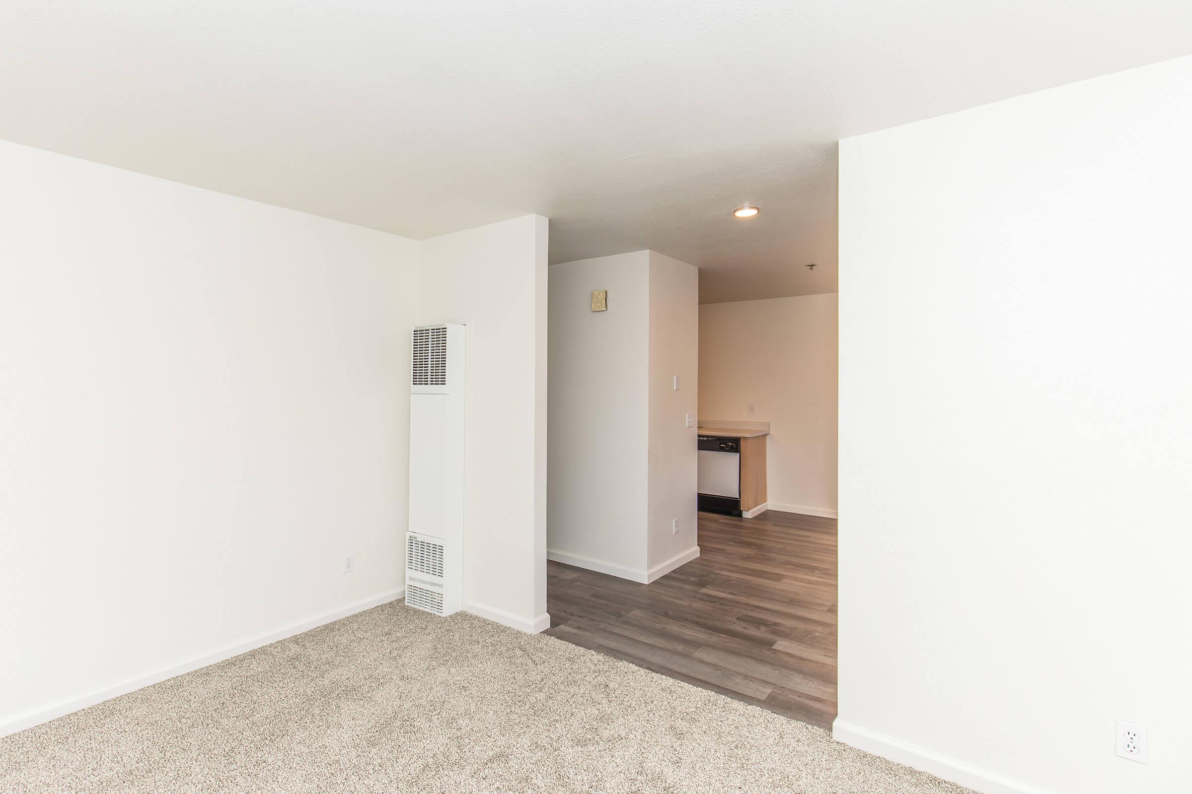 A spacious, empty room with light-colored walls and carpeted flooring. To the left, there is a wall-mounted heater. In the background, a doorway leads to another area featuring a kitchenette. The overall setting is bright and inviting, suggesting potential for decoration and personalization.