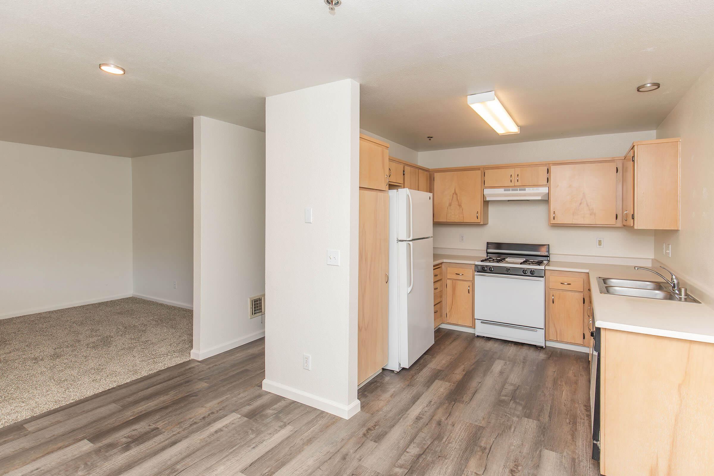 A spacious kitchen with light wood cabinetry, a white refrigerator, gas stove, and double sink. The floor is laminate, and there is a carpeted area visible in the adjacent room. Soft lighting and neutral wall colors create a bright, open atmosphere.