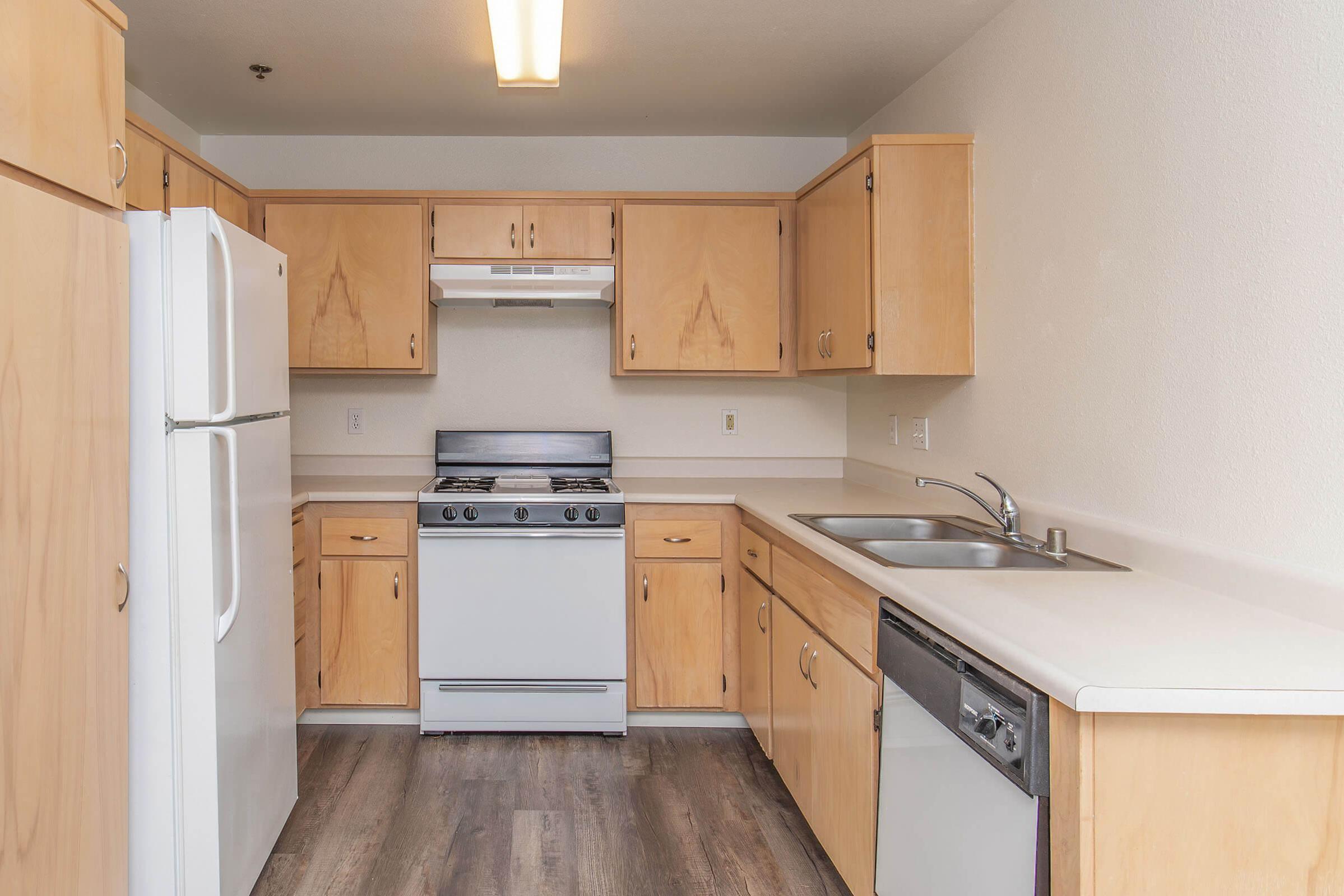 A modern kitchen featuring light wood cabinets, a white refrigerator, a gas stove with a hood, and a stainless steel dishwasher. The countertops are light-colored, and there are dual sinks. The flooring is a wood-like laminate, creating a warm and inviting atmosphere.