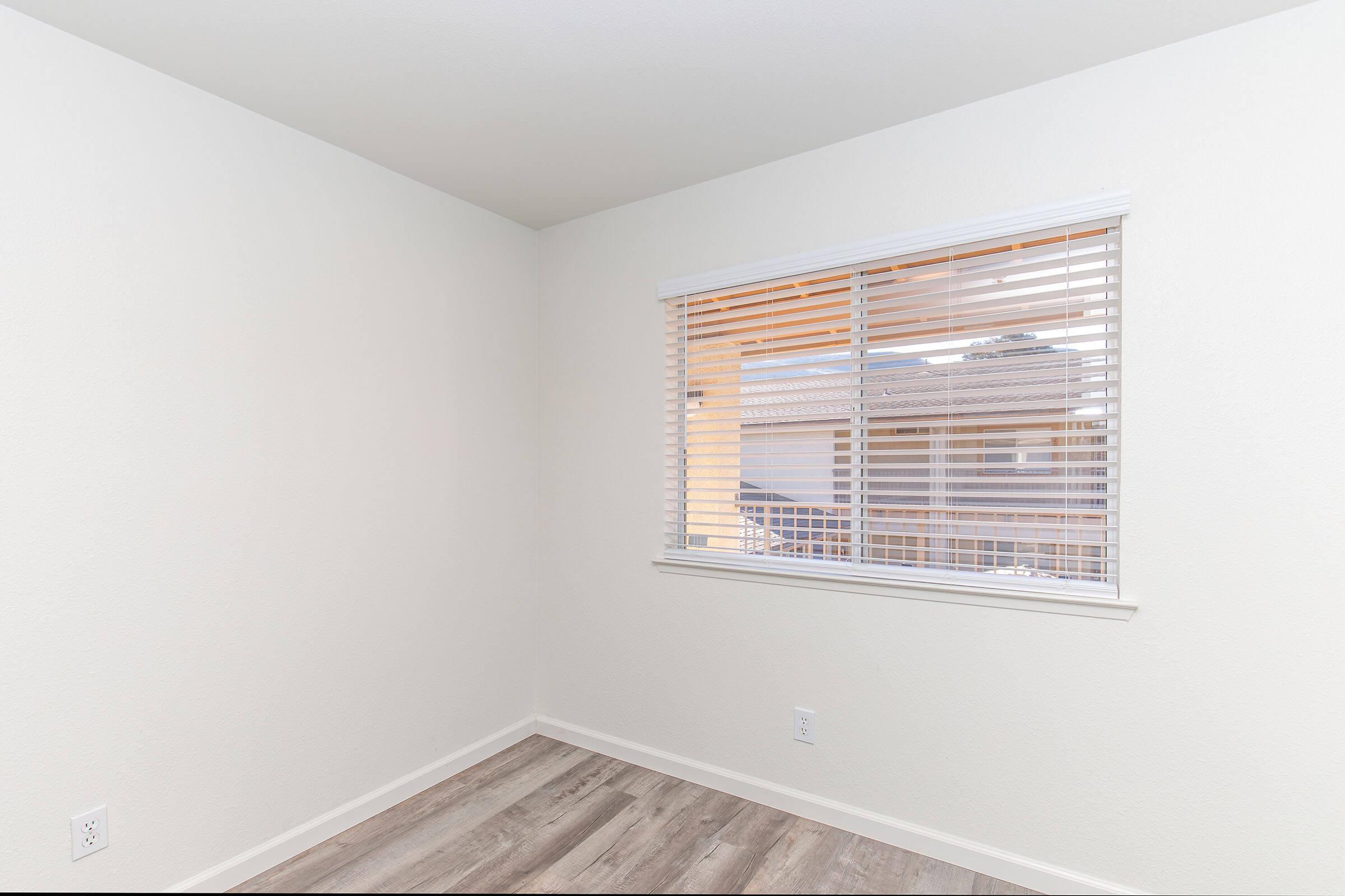 Empty room with light-colored walls and a window covered by white blinds. The floor is light wood, and there is a minimalistic design. The window offers a view of an outdoor area, but details outside are not visible. The overall aesthetic is clean and modern.