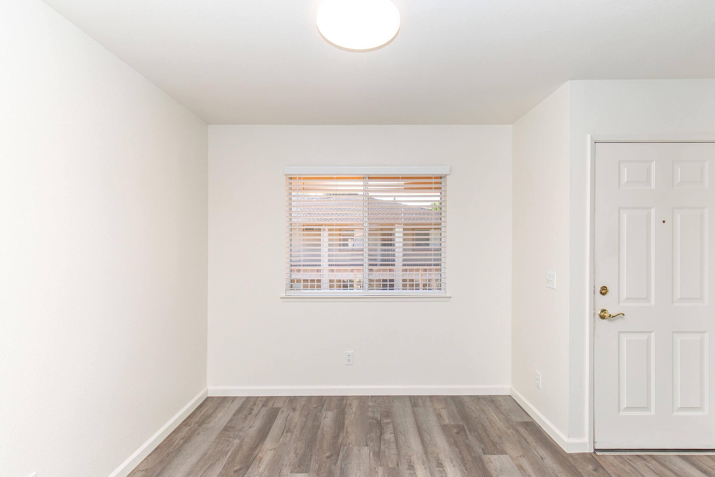 A bright, empty room with light-colored walls and a window with blinds. The flooring is a sleek, wood-like laminate. A door with a brass handle is visible on the right side, contributing to the minimalist and modern aesthetic of the space. Ideal for various uses or decoration.