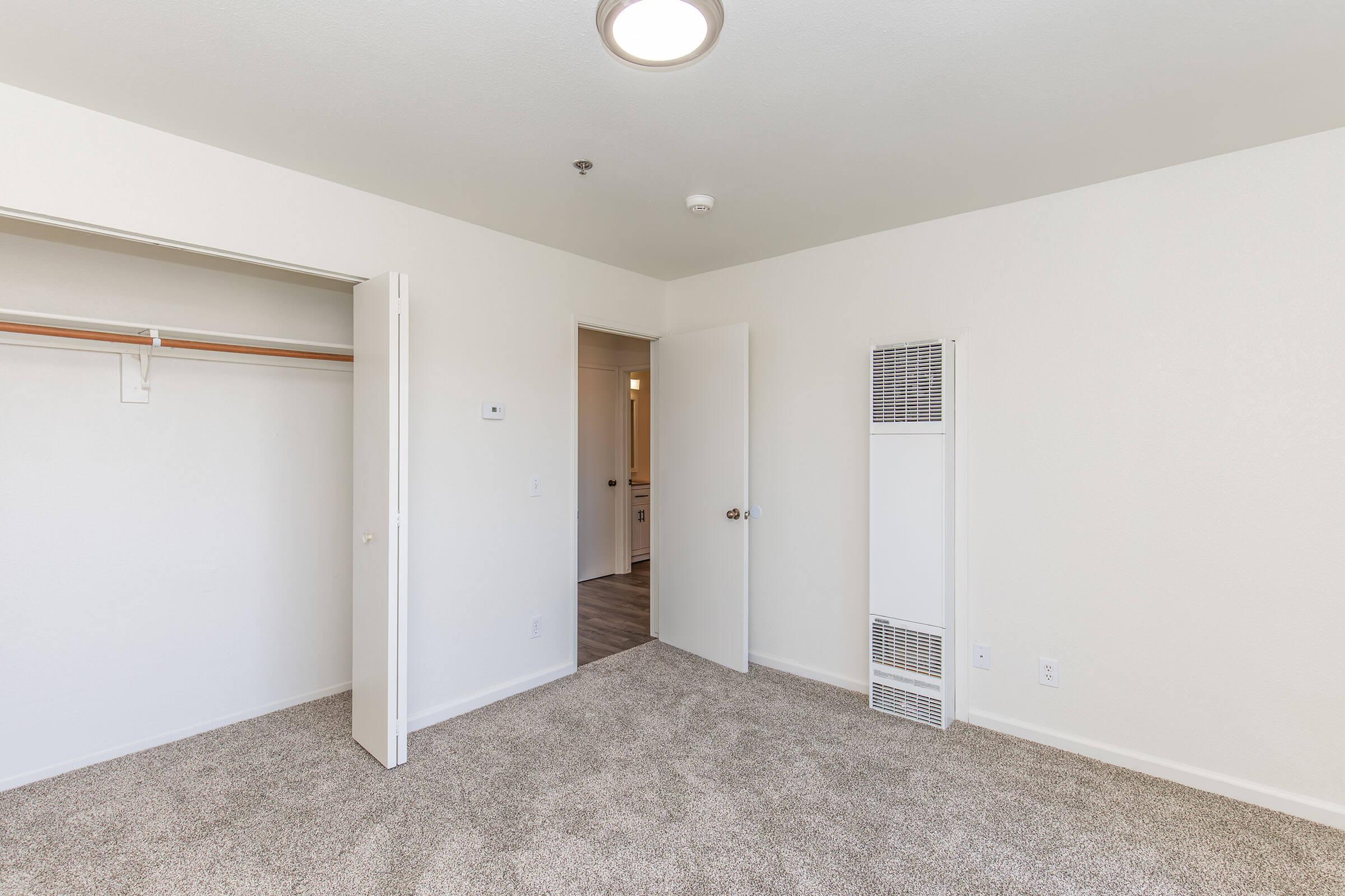 A spacious, empty room with light beige carpet and white walls. It features a closet with sliding doors on the left, a door to another room straight ahead, and a wall-mounted heater on the right. The ceiling is bright with a round light fixture, creating a clean and inviting ambiance.